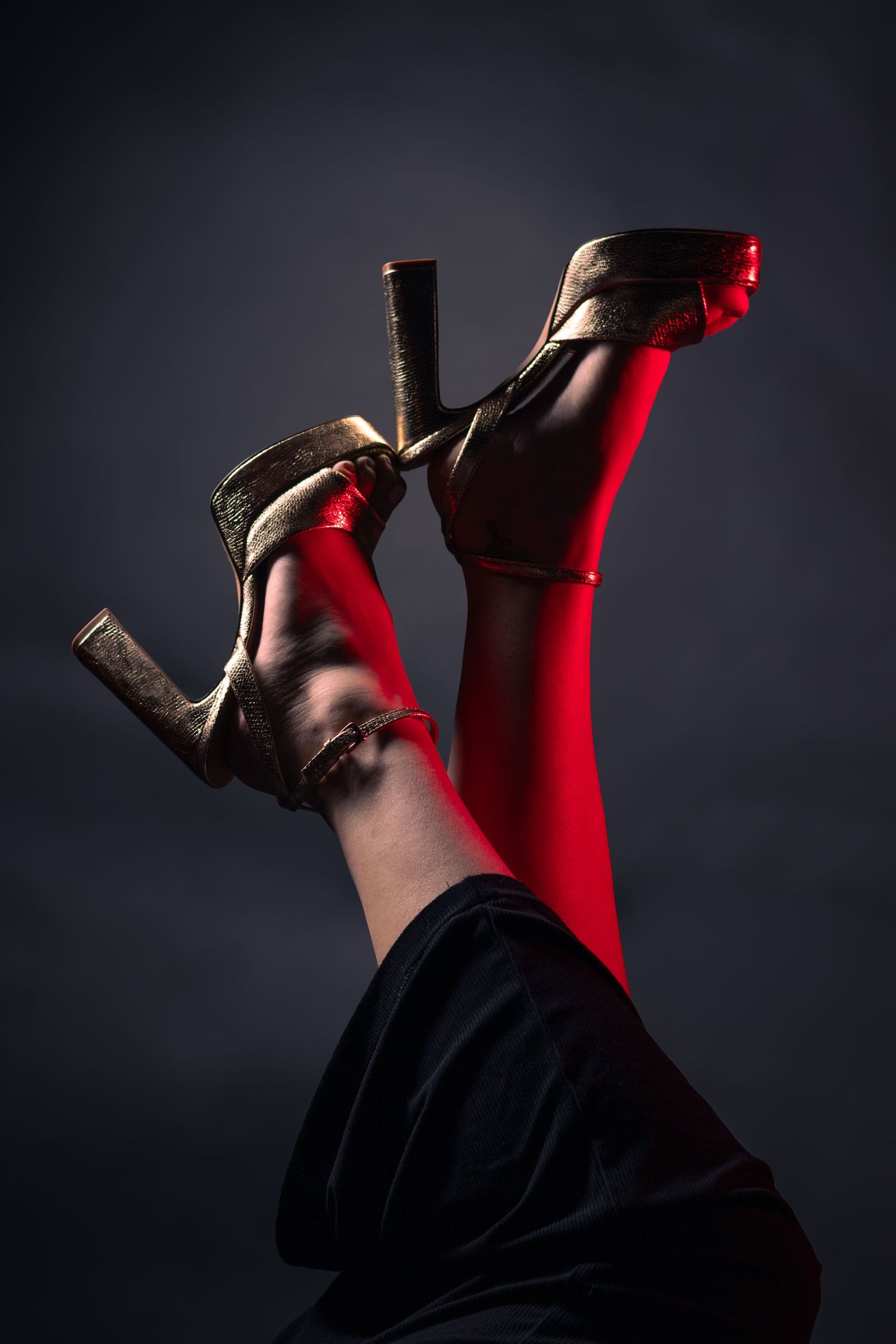 Legs raised up wearing gold platform heels and red stockings against dark background.
