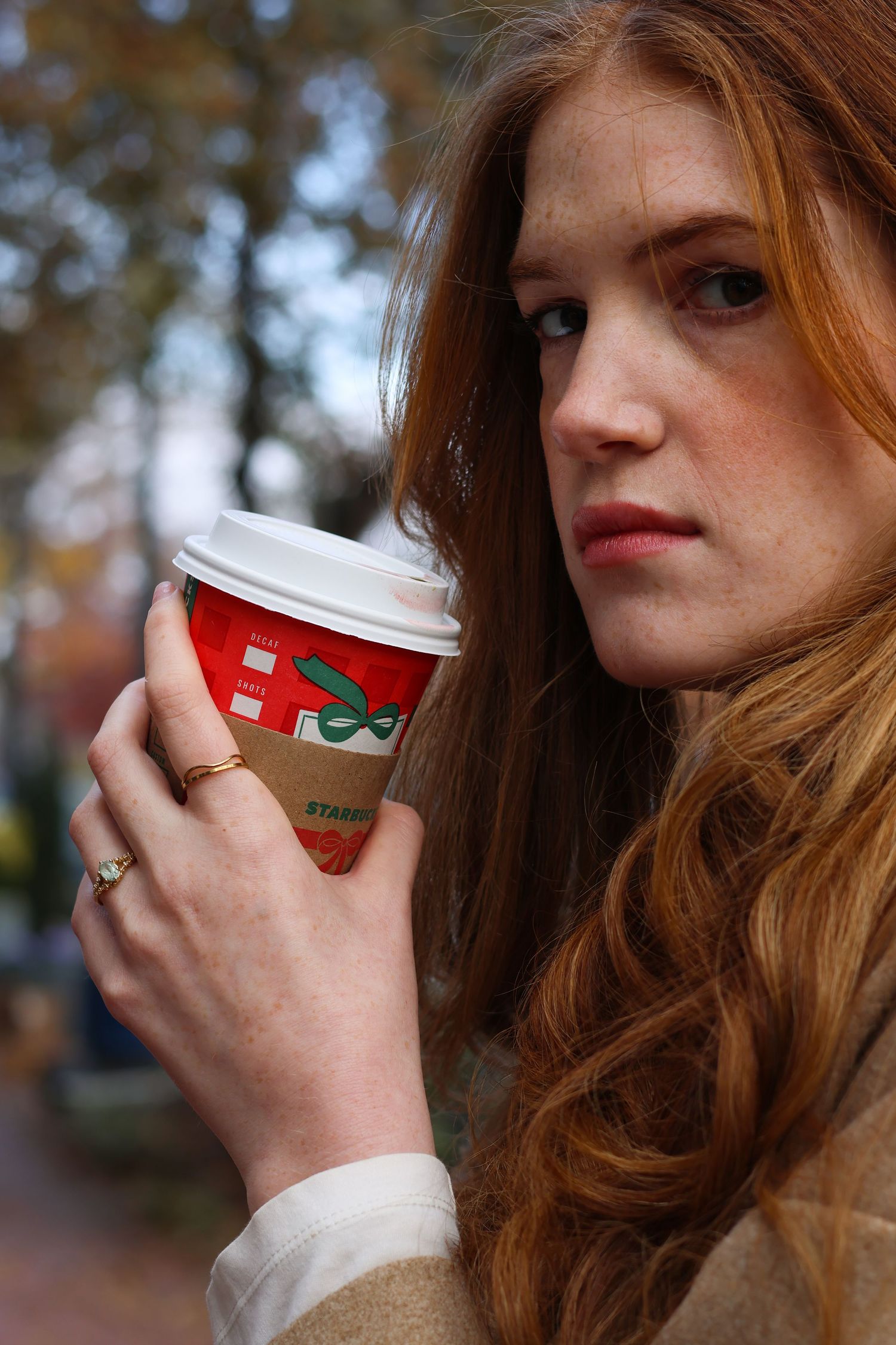 Young redhead woman with freckles holding a Starbucks holiday cup outdoors, wearing a beige coat and gold rings.