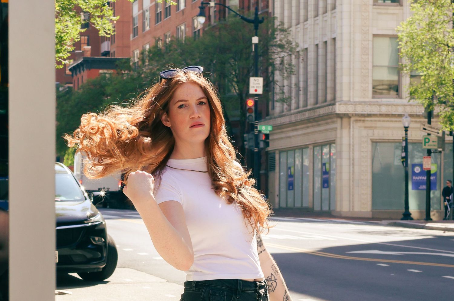Young woman with flowing red hair in white top poses on sunny urban street corner with city buildings behind her.