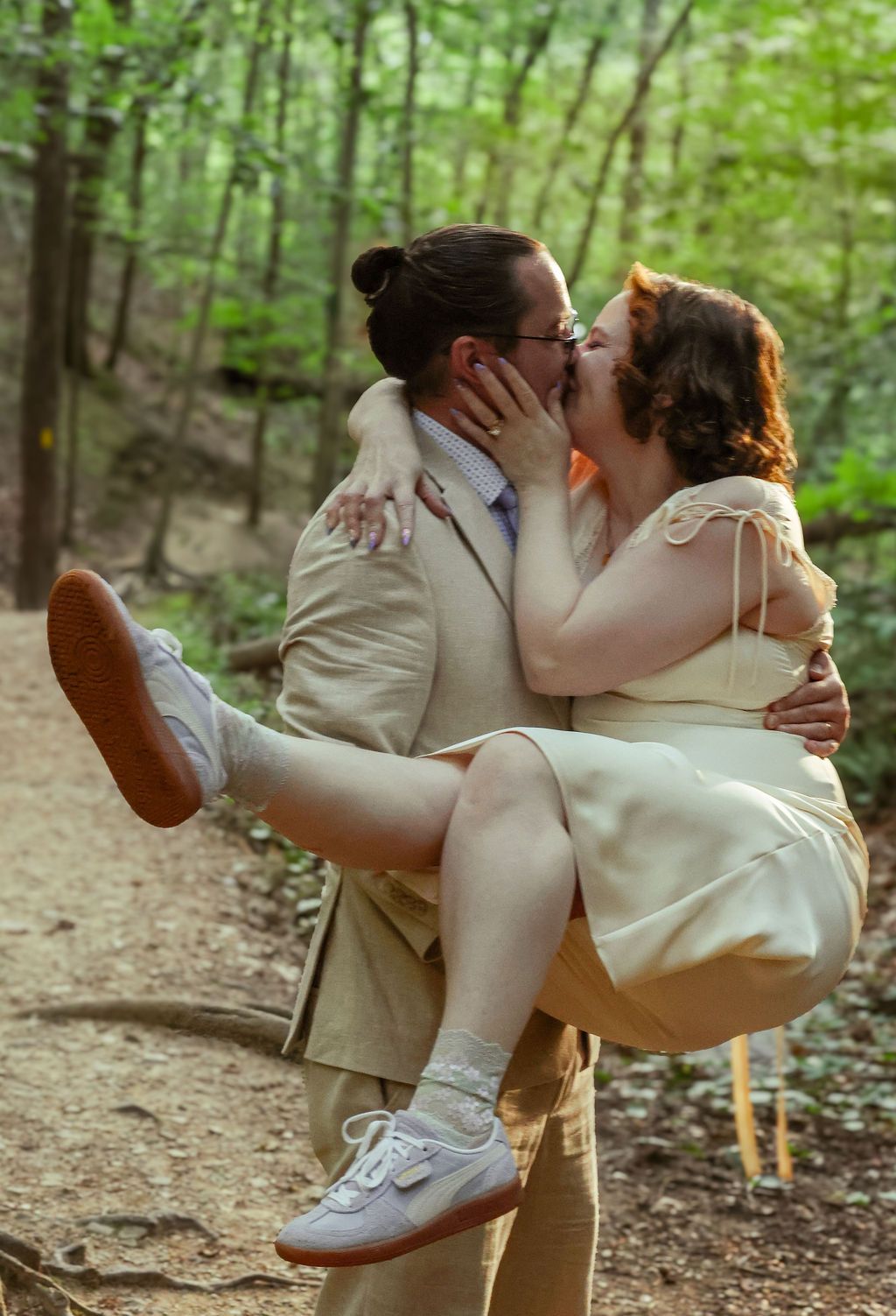 A man in a tan suit lifts a woman in a cream dress off the ground as they kiss on a sunlit wooded forest trail.