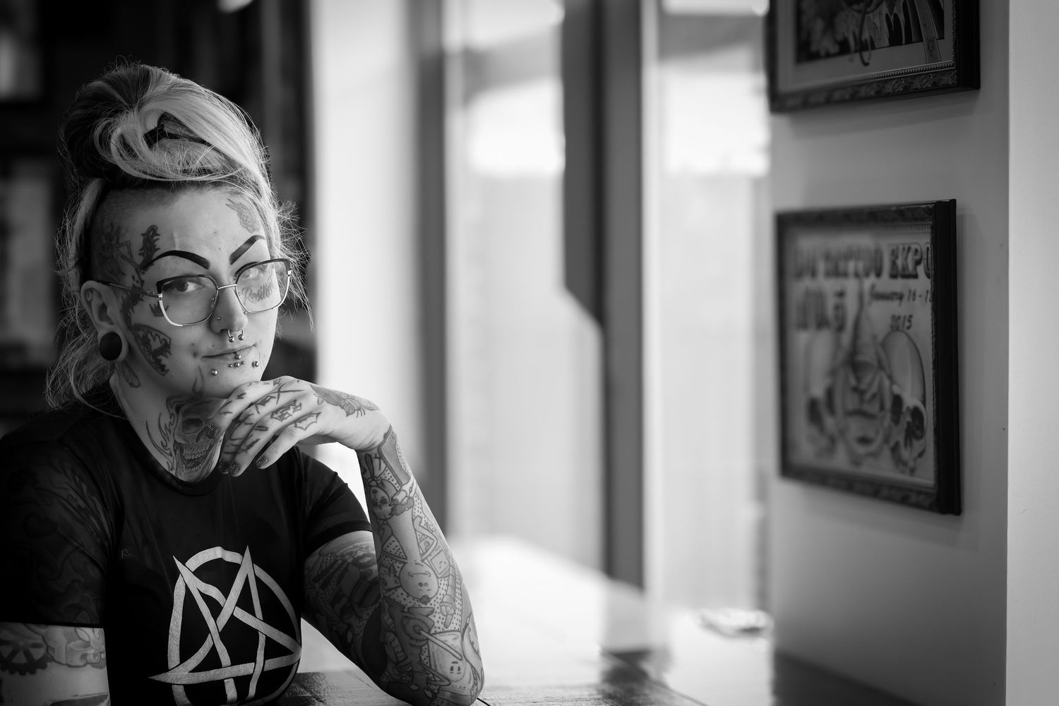 Black and white portrait of someone with tattoos and glasses sitting thoughtfully in front of framed wall art.