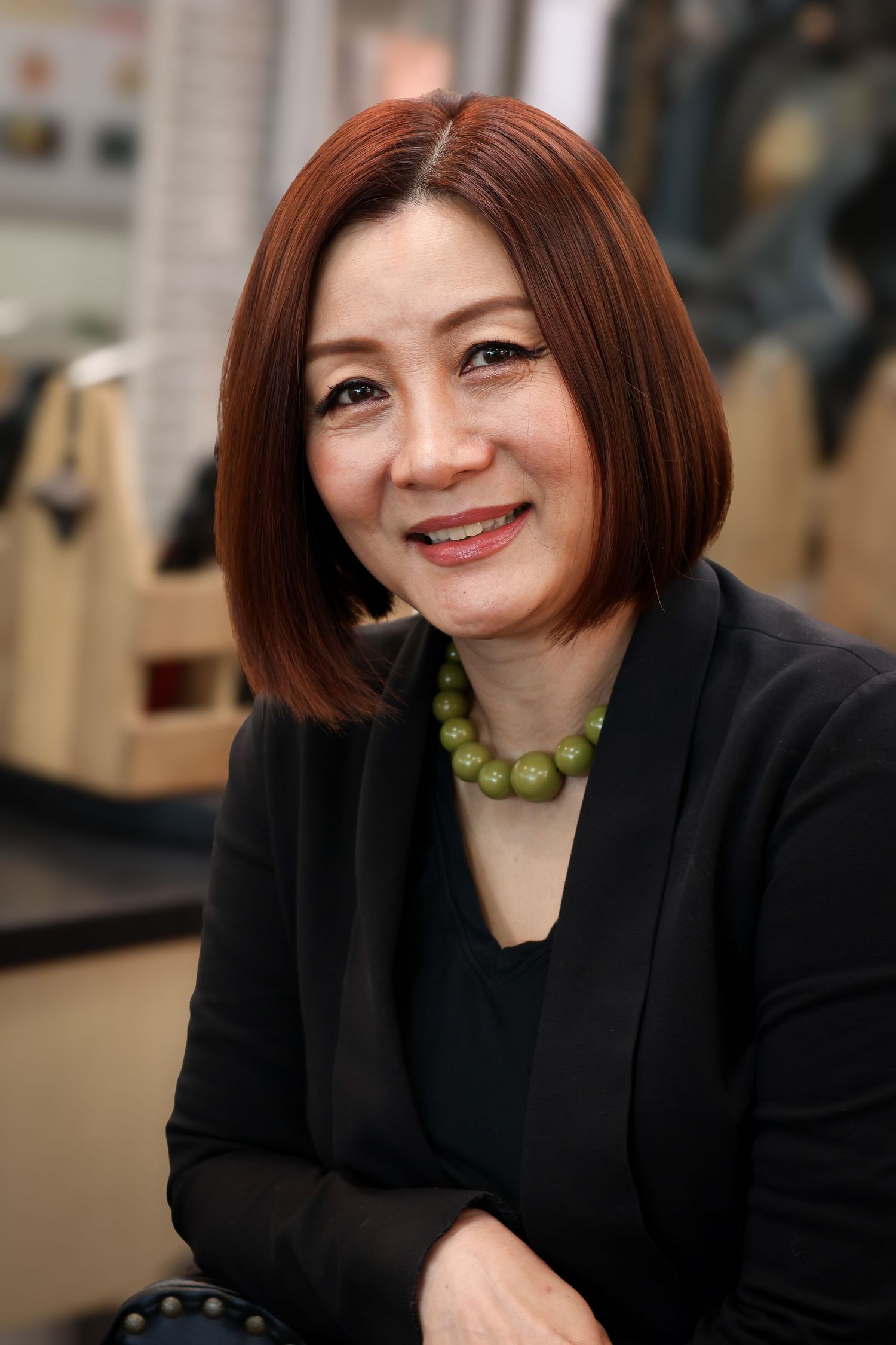 Professional portrait of someone in a black blazer and green beaded necklace with short auburn hair smiling warmly.