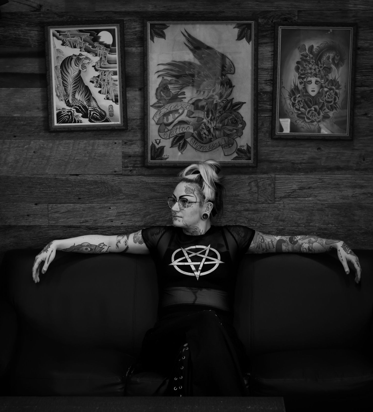Black and white photo of someone in alternative style clothing seated against a wall with framed artwork displayed above.