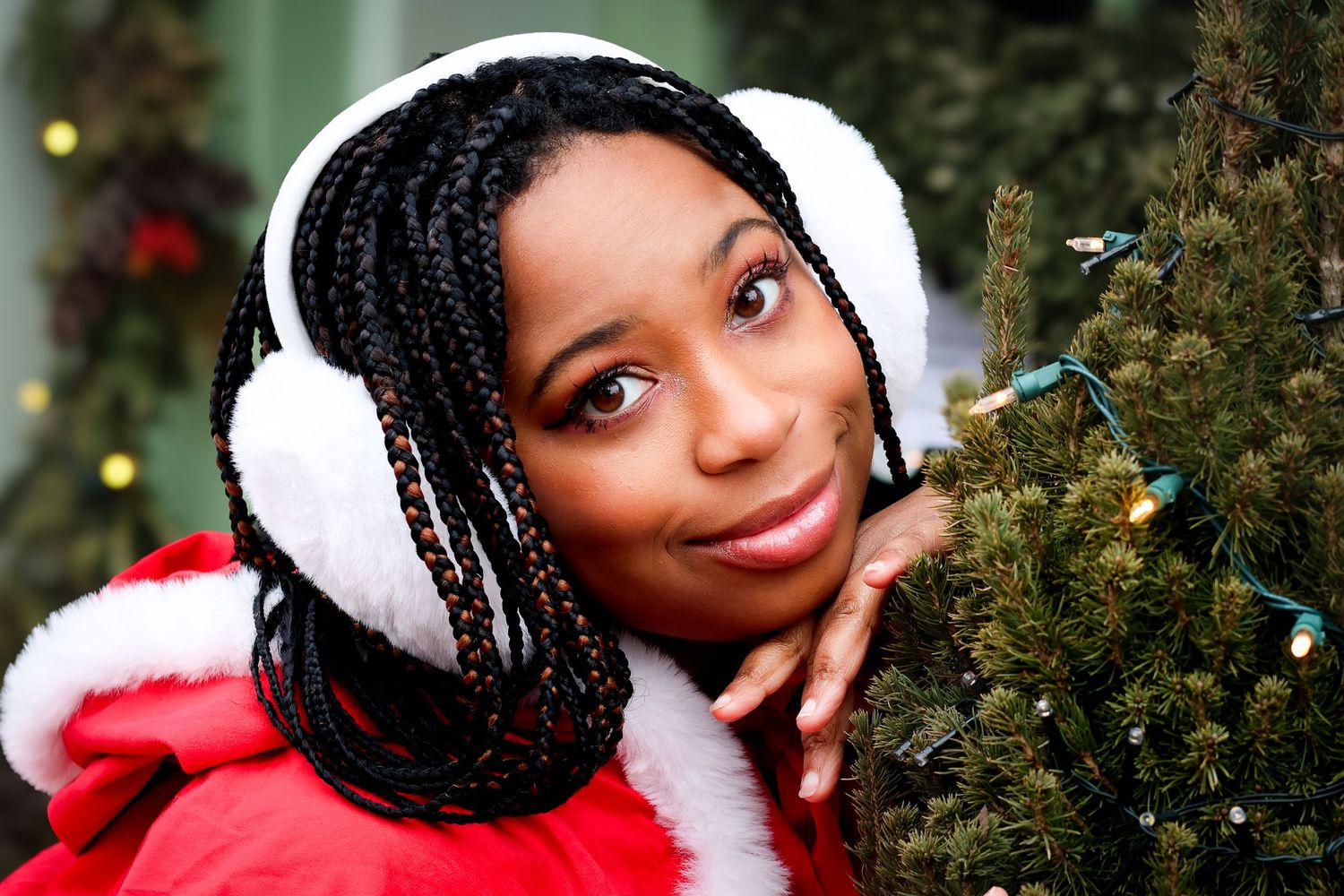 Young woman in Santa outfit with white earmuffs and braids posing beside a decorated Christmas tree.