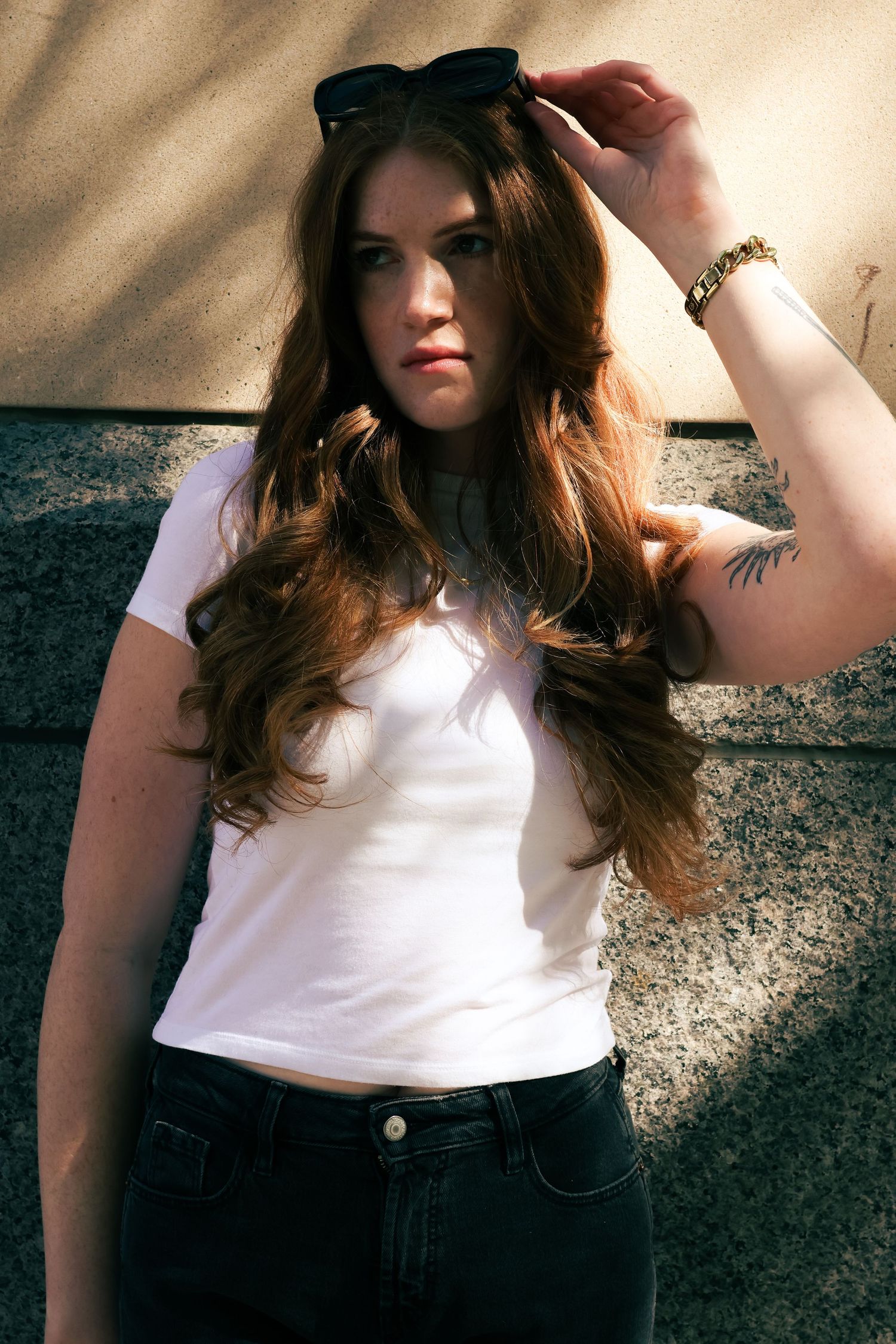 Young woman with long auburn hair in white crop top and black jeans, holding sunglasses, showing arm tattoo.