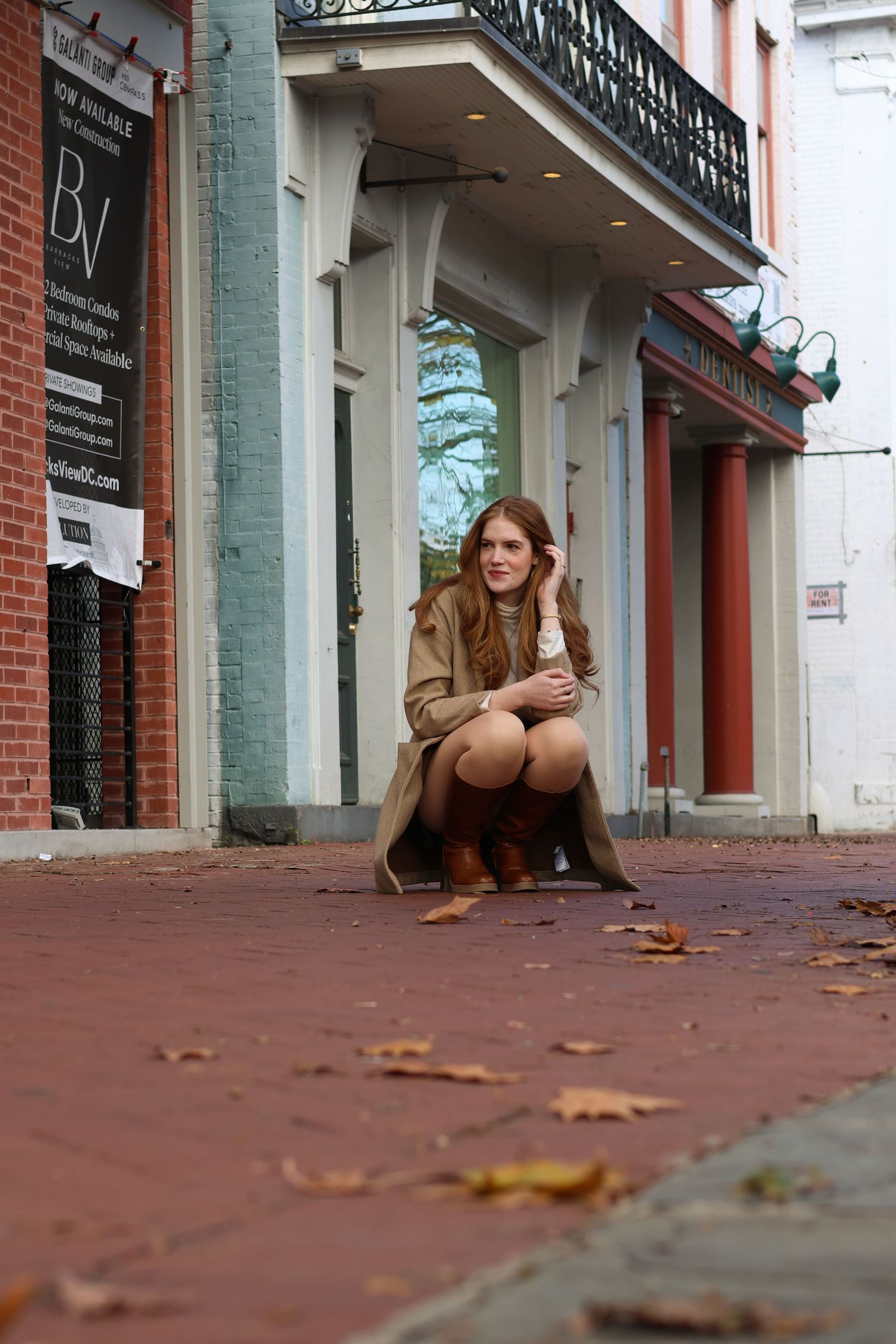 Young woman in tan trench coat crouching on brick sidewalk outside vintage storefronts with fall leaves.