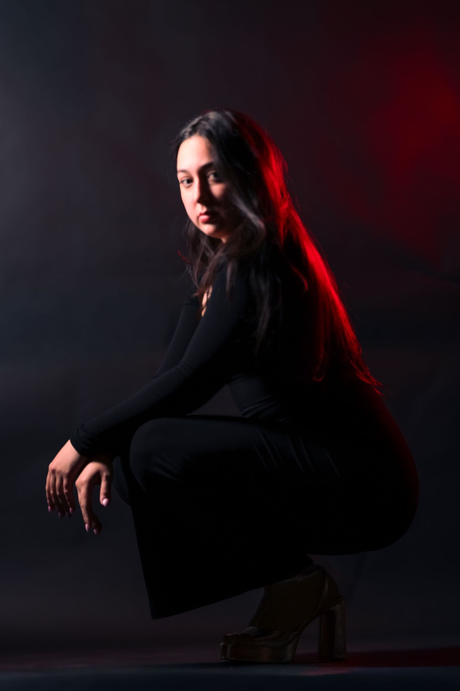 Woman in black outfit squatting gracefully in red and dark studio lighting.