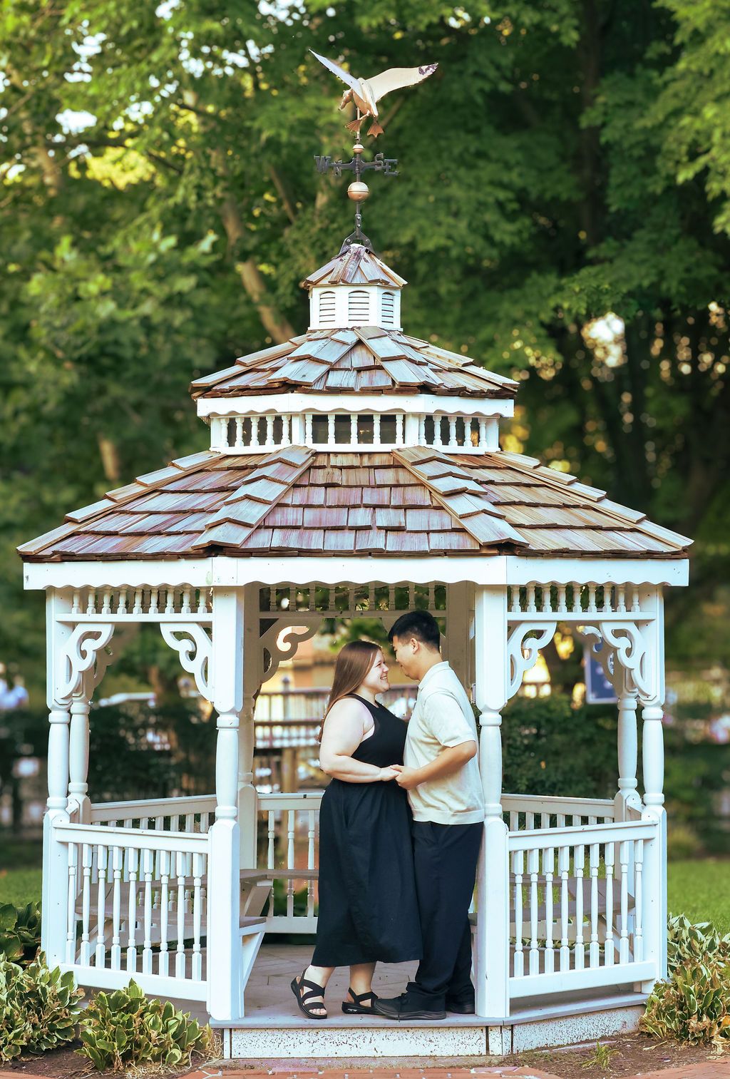 A couple stands close together inside a white wooden gazebo with a shingled roof and bird weathervane, surrounded by lush green trees.