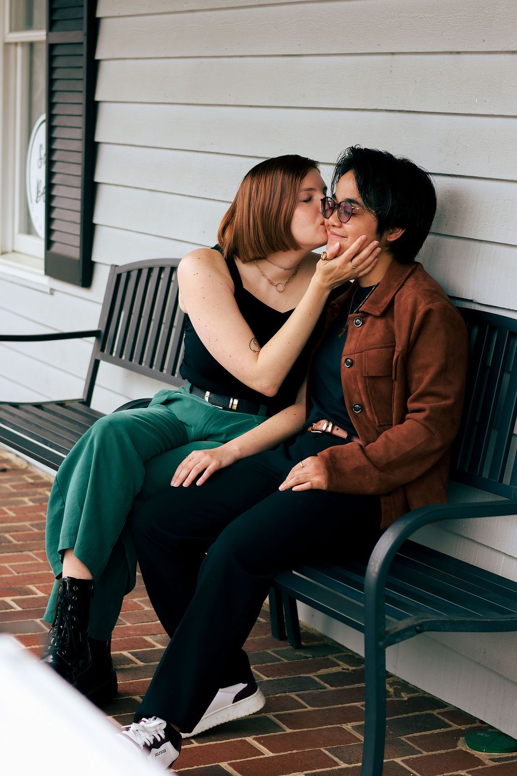 Two people sit close together on a green park bench on a brick patio, one kissing the other's cheek while hugging in a tender moment.