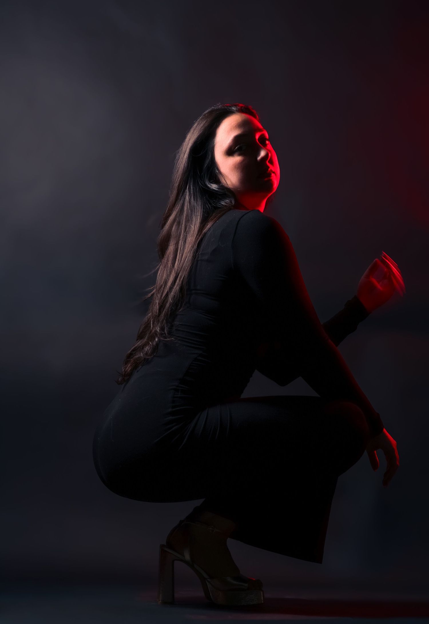 Woman in black outfit crouching with red dramatic lighting against dark studio background.