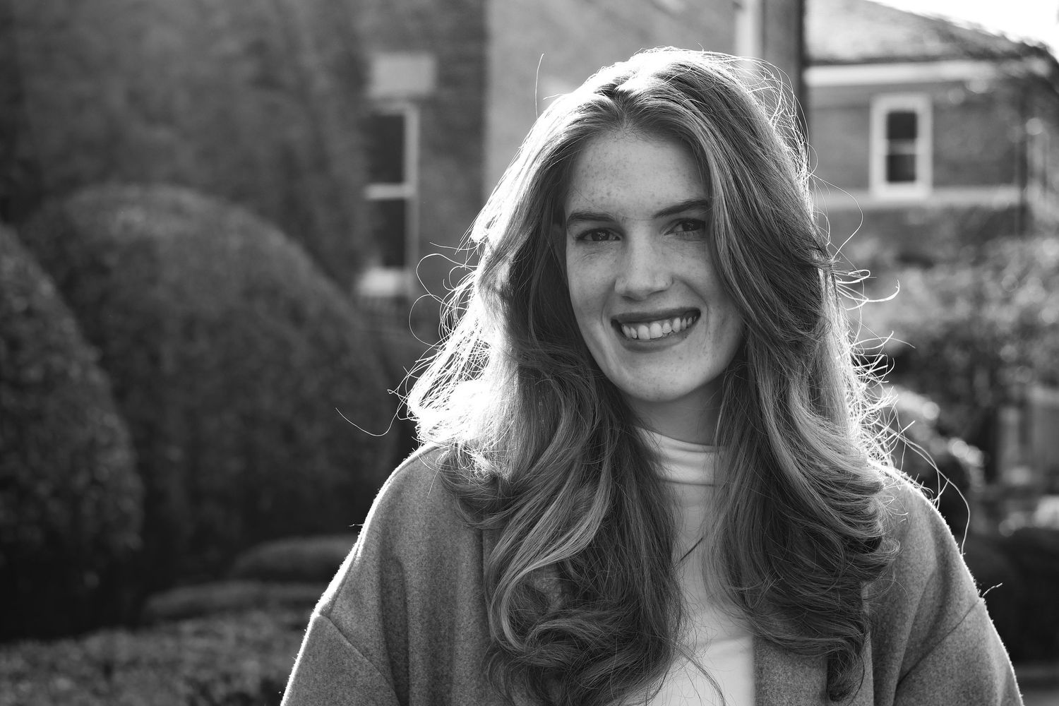 Black and white portrait of a smiling young woman with long wavy hair outdoors near hedges.