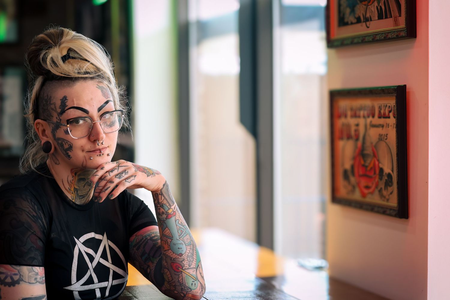 A person with tattoos and glasses sits contemplatively in a room with framed artwork on the walls.