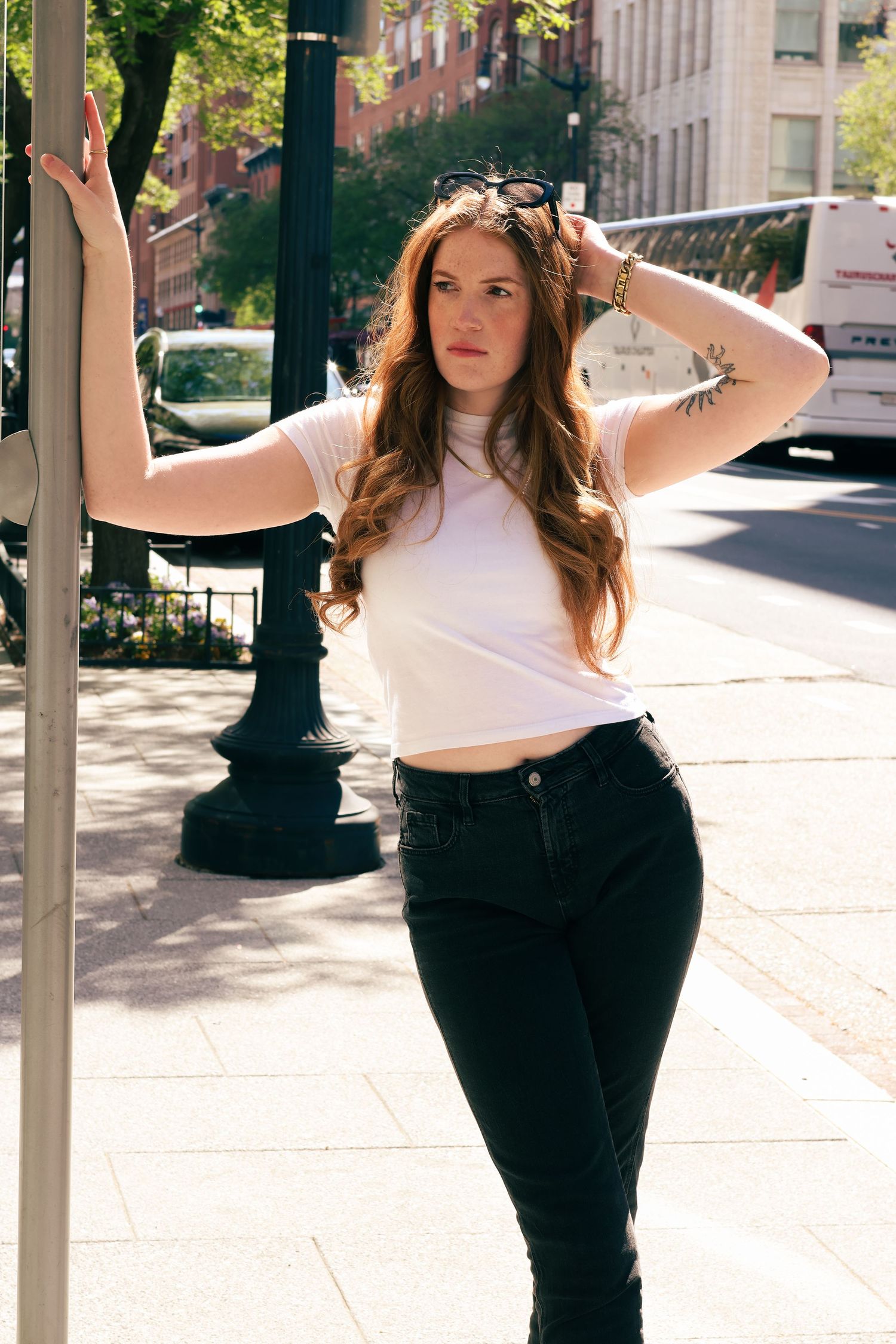 Young woman with long auburn hair in white crop top and black jeans poses by a street pole in an urban setting.