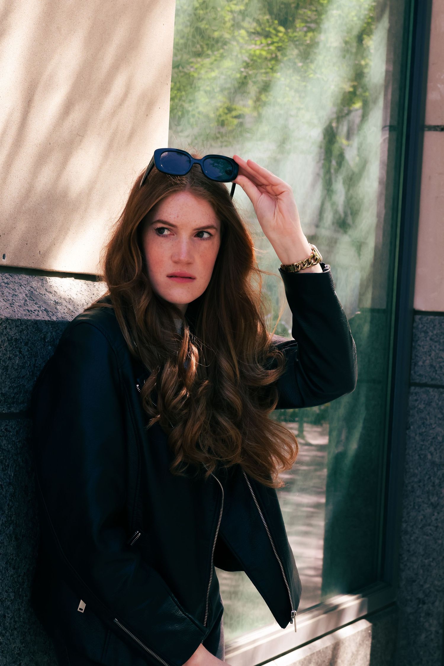 Young woman with long wavy hair holding sunglasses on head, wearing black jacket, leaning against wall outdoors.