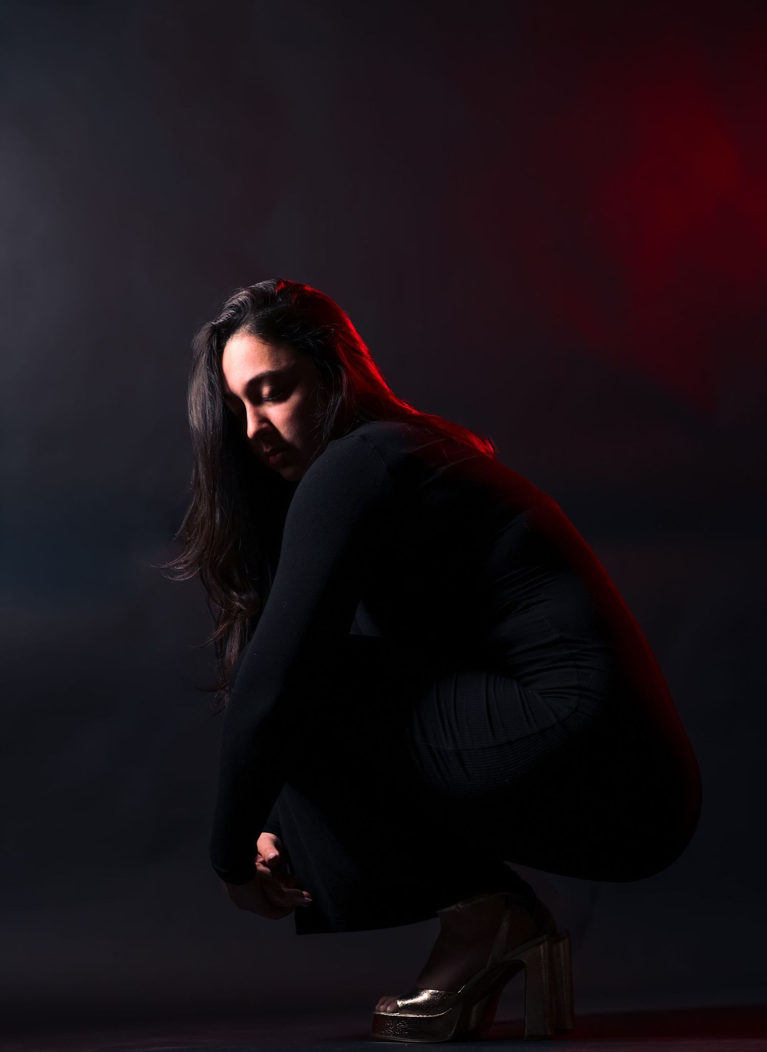 Woman in black outfit crouches gracefully with red dramatic lighting in dark studio setting.