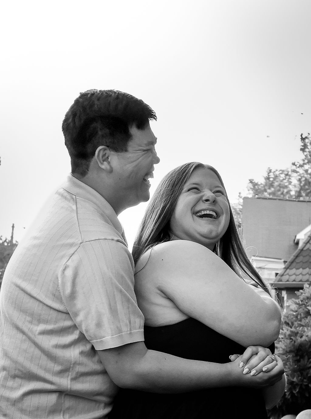 Black and white photo of a man and woman embracing outdoors as they laugh together during an engagement photo session.