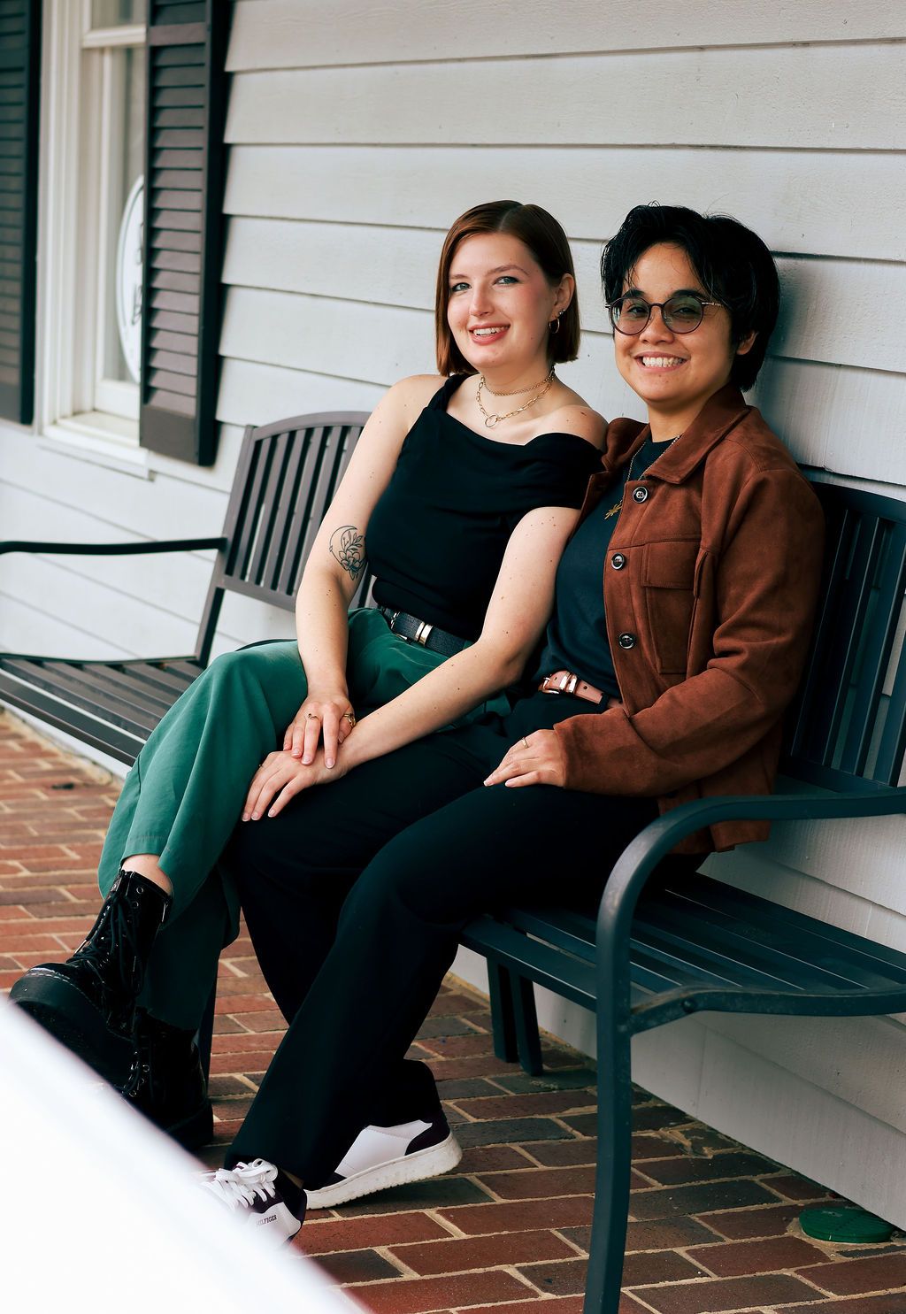 Two smiling women sit close together on a dark green bench on a brick porch, one in a black top and green pants, the other in a rust jacket.
