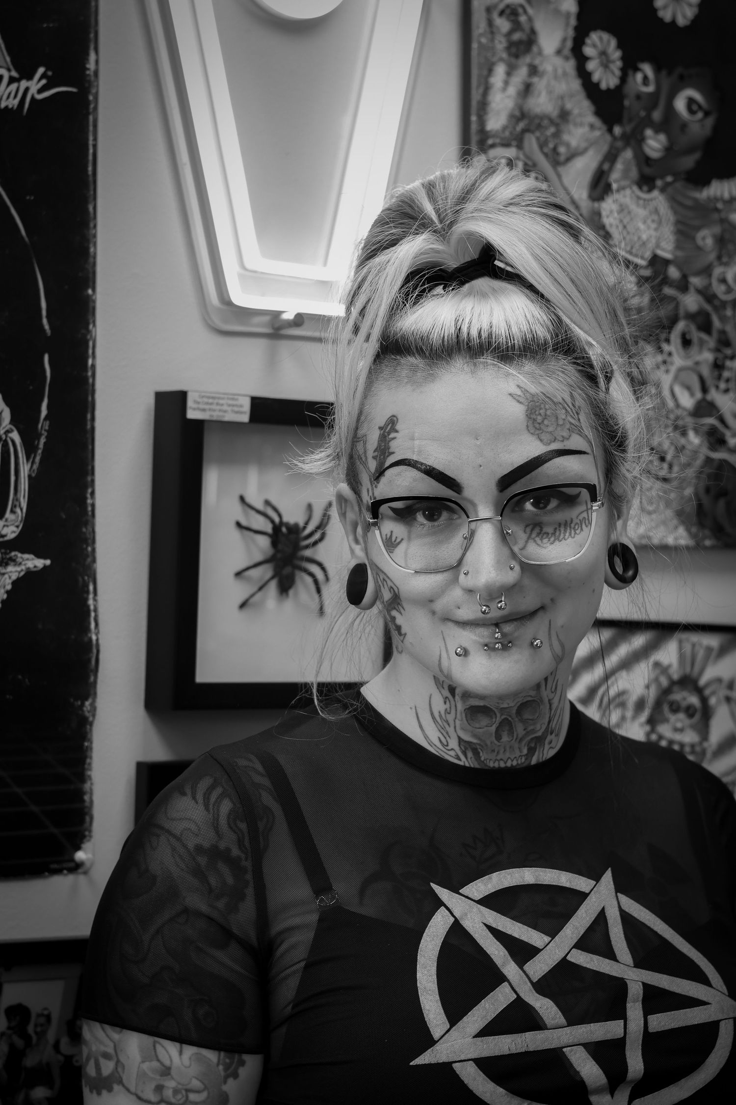Black and white portrait of someone wearing glasses and a pentagram shirt with visible tattoos and piercings.