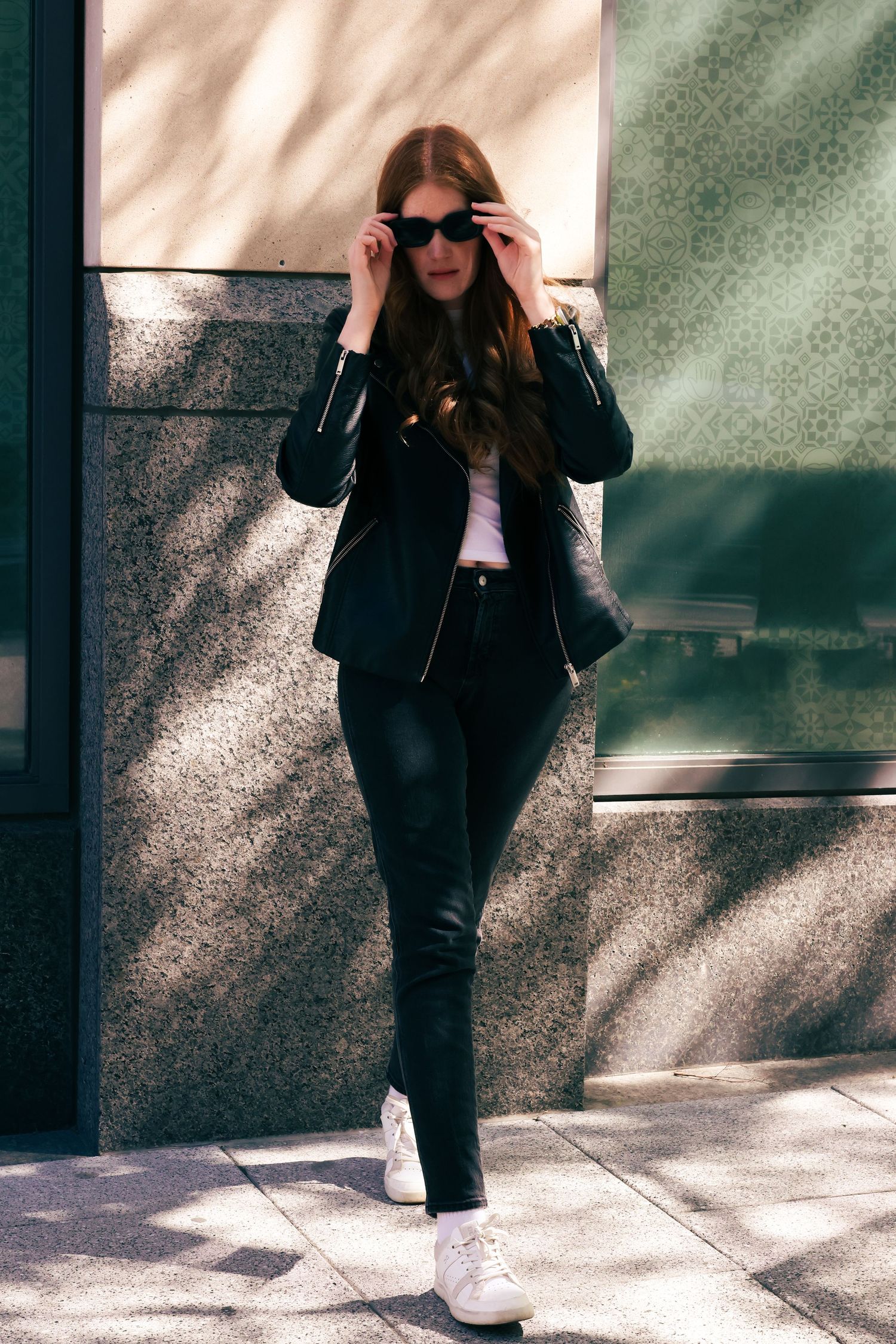 Stylish woman in black leather jacket, pants, and white sneakers adjusting sunglasses on city sidewalk.
