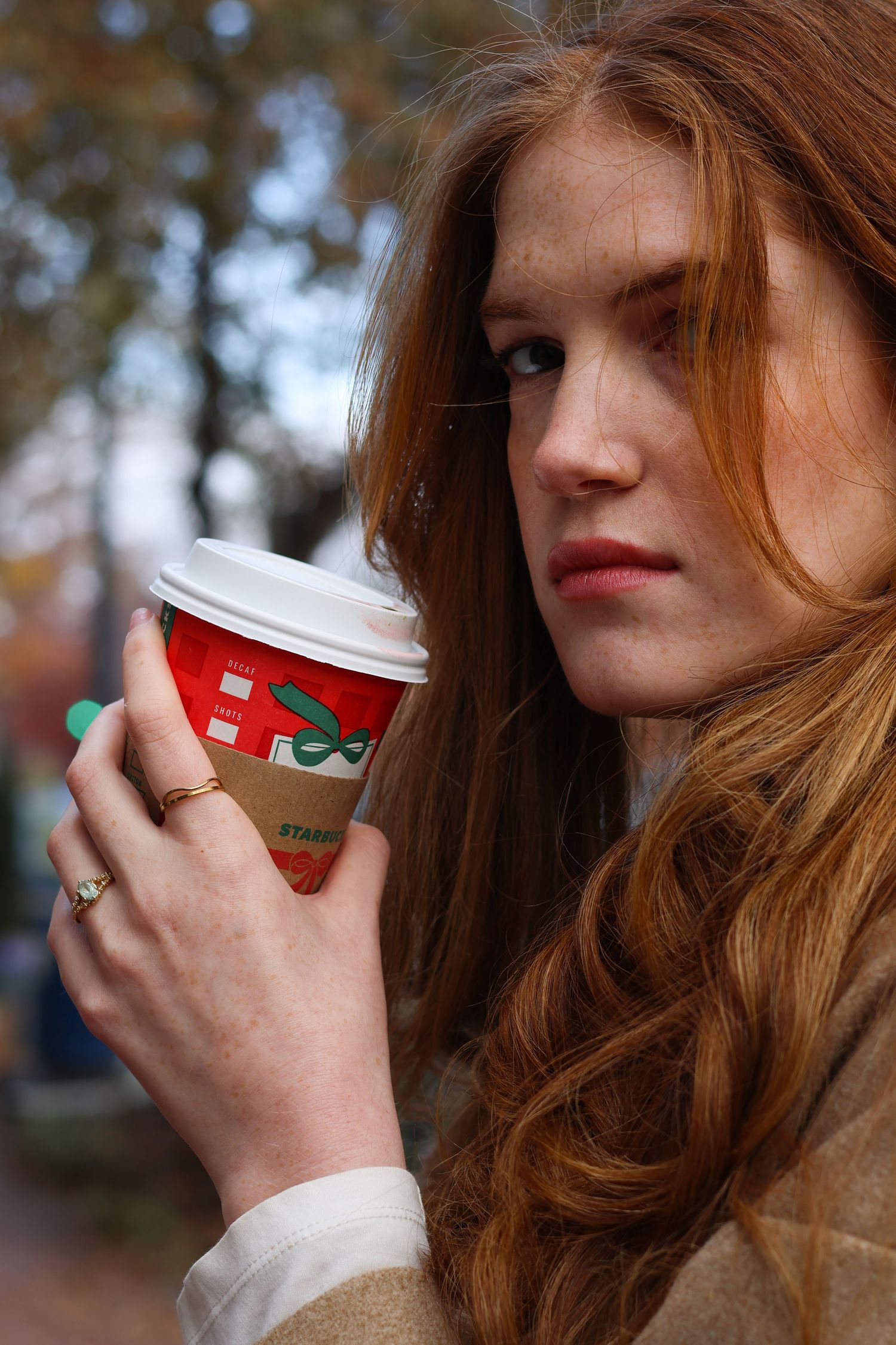 Red-haired woman holding a Starbucks holiday cup outdoors in autumn.