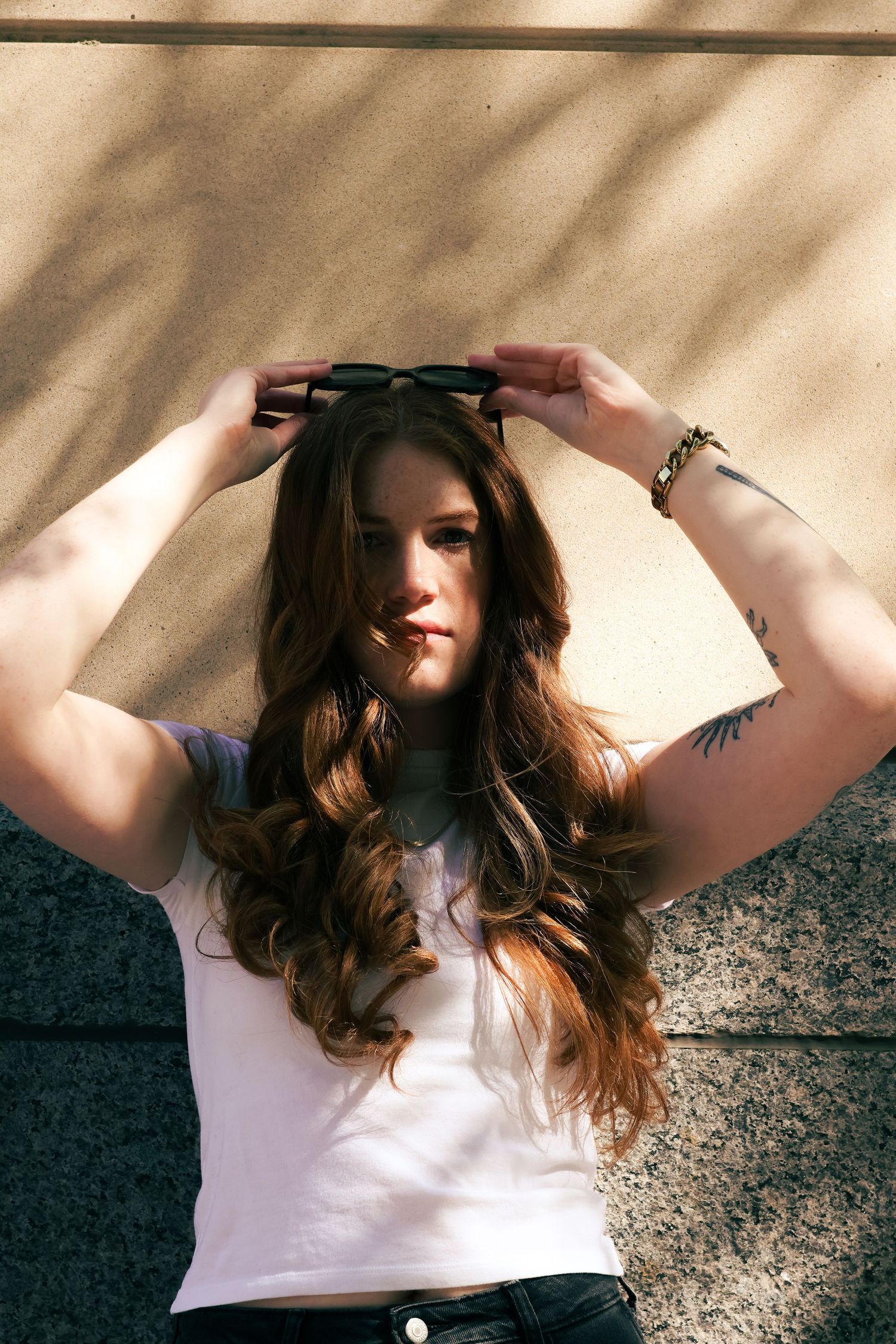 Young woman with long wavy auburn hair adjusting sunglasses on head, wearing white crop top, tattoo visible on arm.