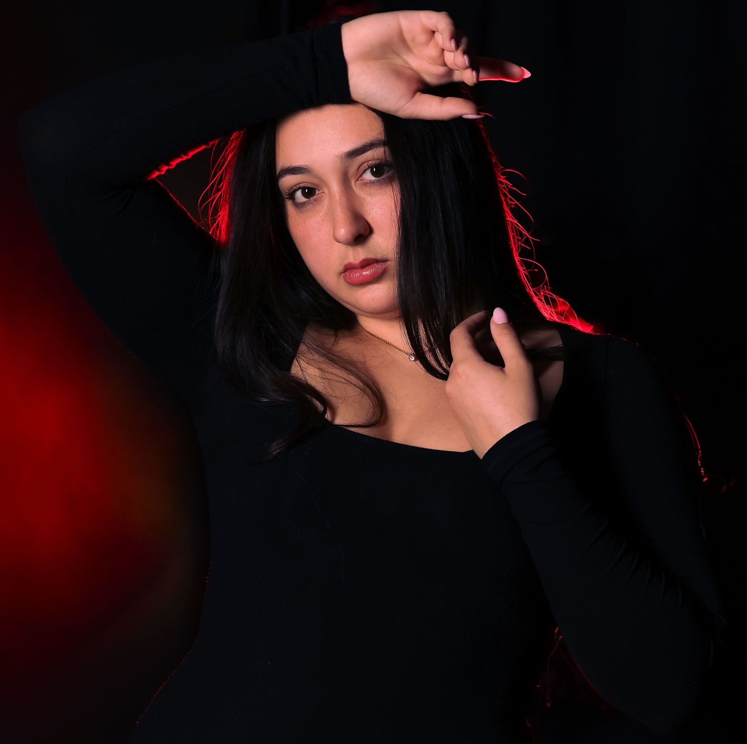 Young woman in black outfit poses dramatically with red lighting accents against dark background.