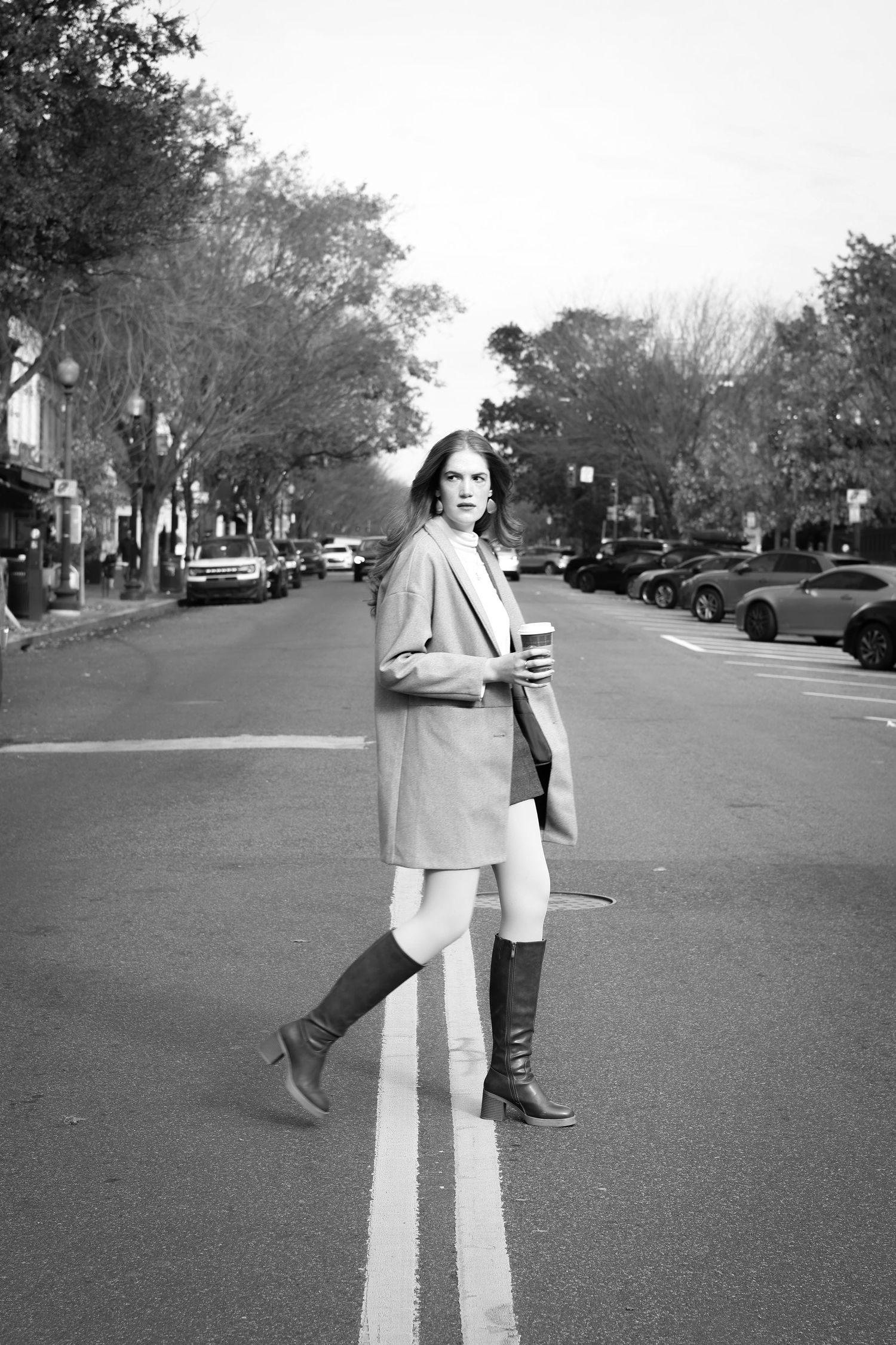 Black and white photo of a woman in a coat and knee-high boots crossing a city street while holding a coffee cup.