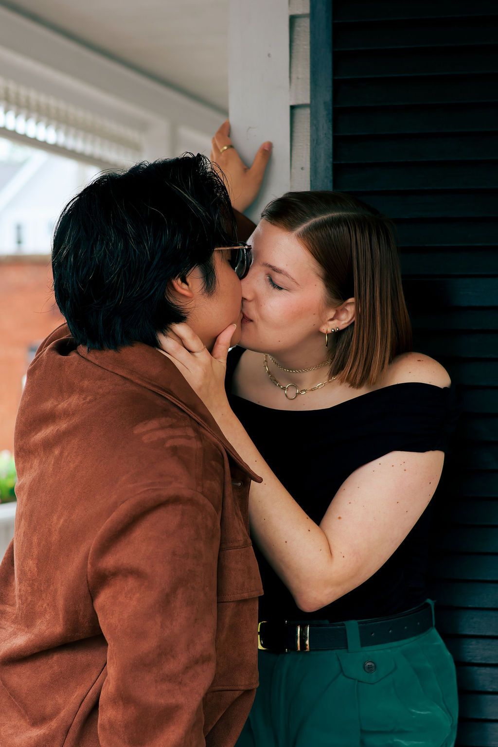 A couple kisses tenderly on a porch, one in a rust-brown jacket and glasses, the other in a black off-shoulder top and teal pants.