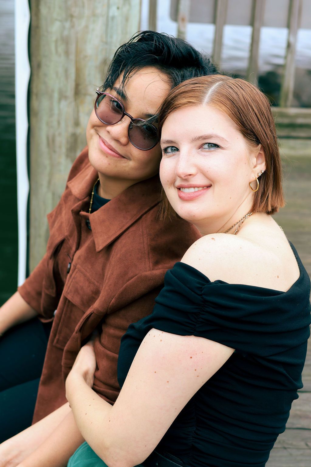 Two smiling women embrace outdoors on a wooden deck, one in a rust-colored shirt and sunglasses, the other in an off-shoulder black top.