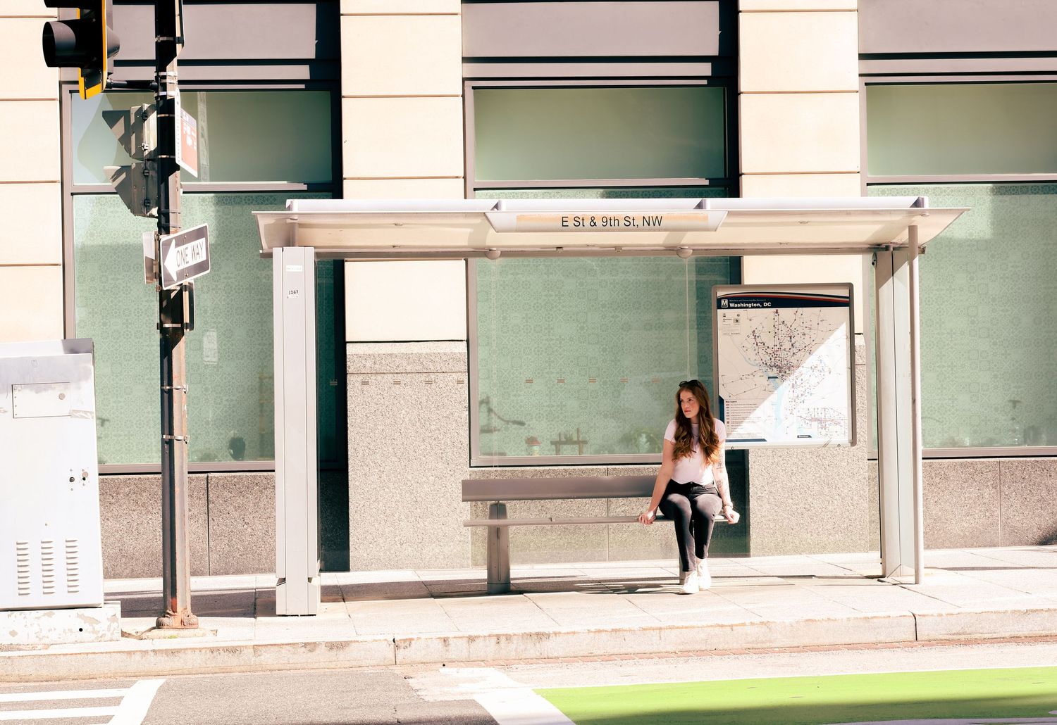 Woman sitting alone at a modern urban bus stop shelter on a sunny city street corner.