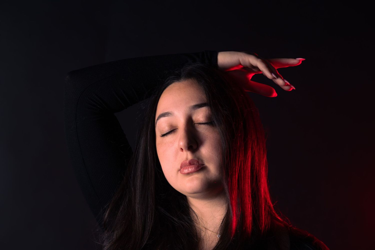 Woman with red-lit hair posing with hand raised above head against dark background.
