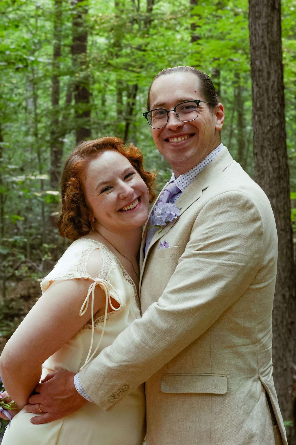 Smiling couple embracing in a sunlit forest, the woman in a cream dress and the man in a tan suit with glasses and a lavender tie.