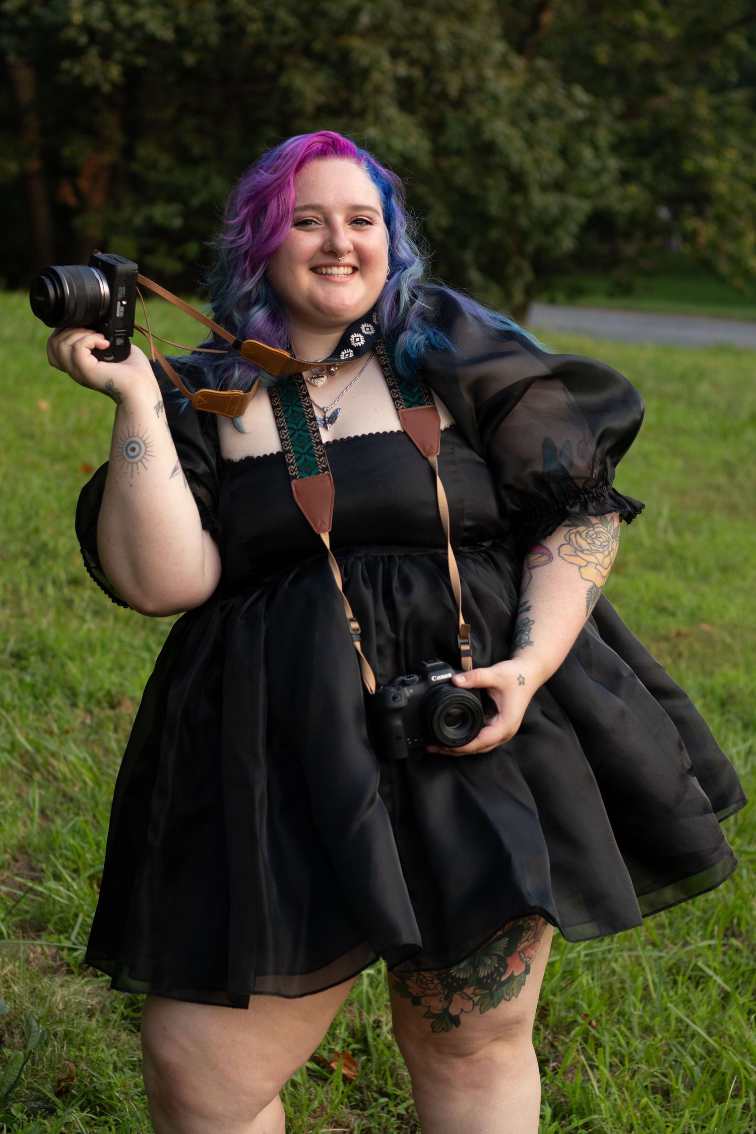 A photographer in a black dress holds a camera while posing outdoors in a grassy field.