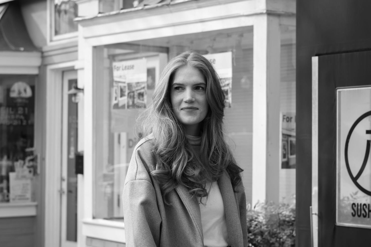 Black and white portrait of someone with long dark hair standing outside a storefront.