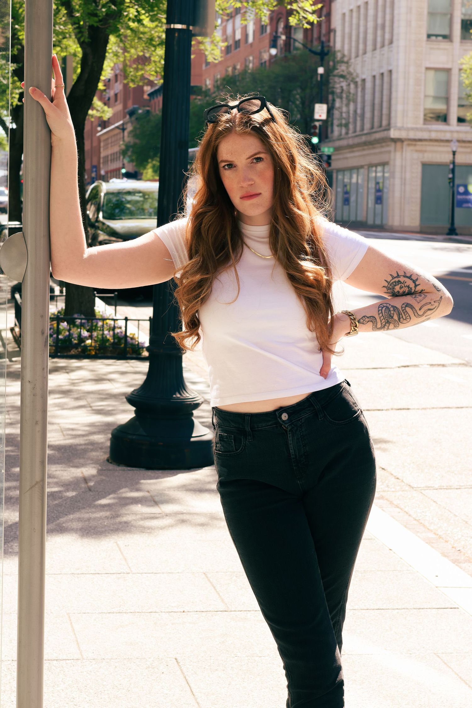 Young woman with long red hair, tattoos, white crop top, and black jeans posing on a sunny urban sidewalk.
