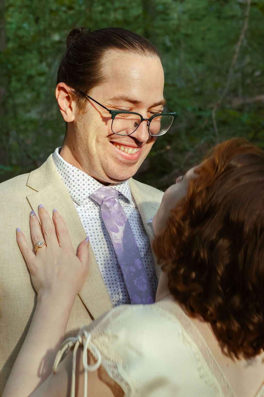 A smiling bearded groom in glasses, tan suit and lavender tie gazes at his bride, whose ringed hand rests on his chest.