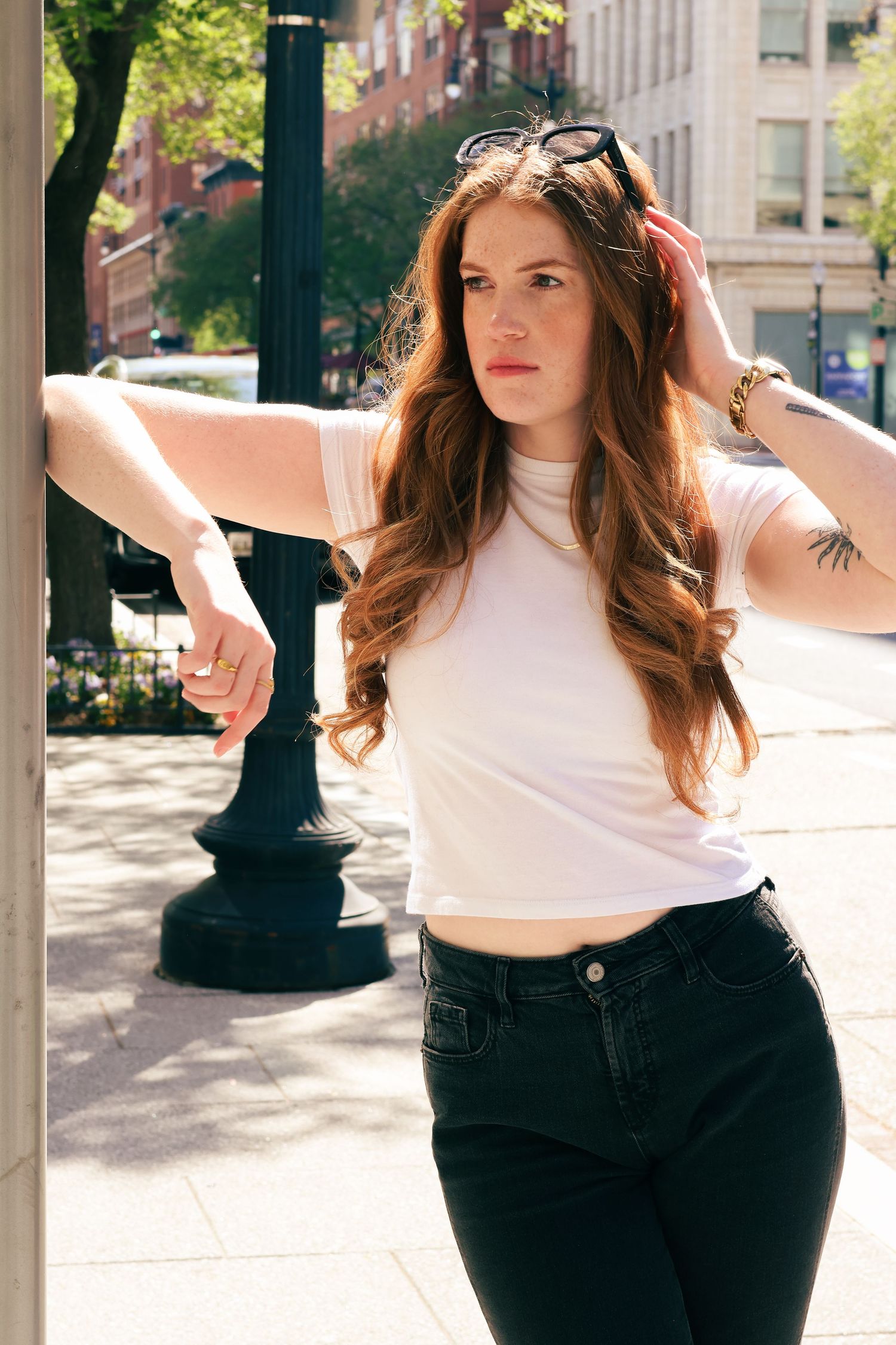 Redhead woman in white crop tee and black jeans leaning on pole on sunny city street.