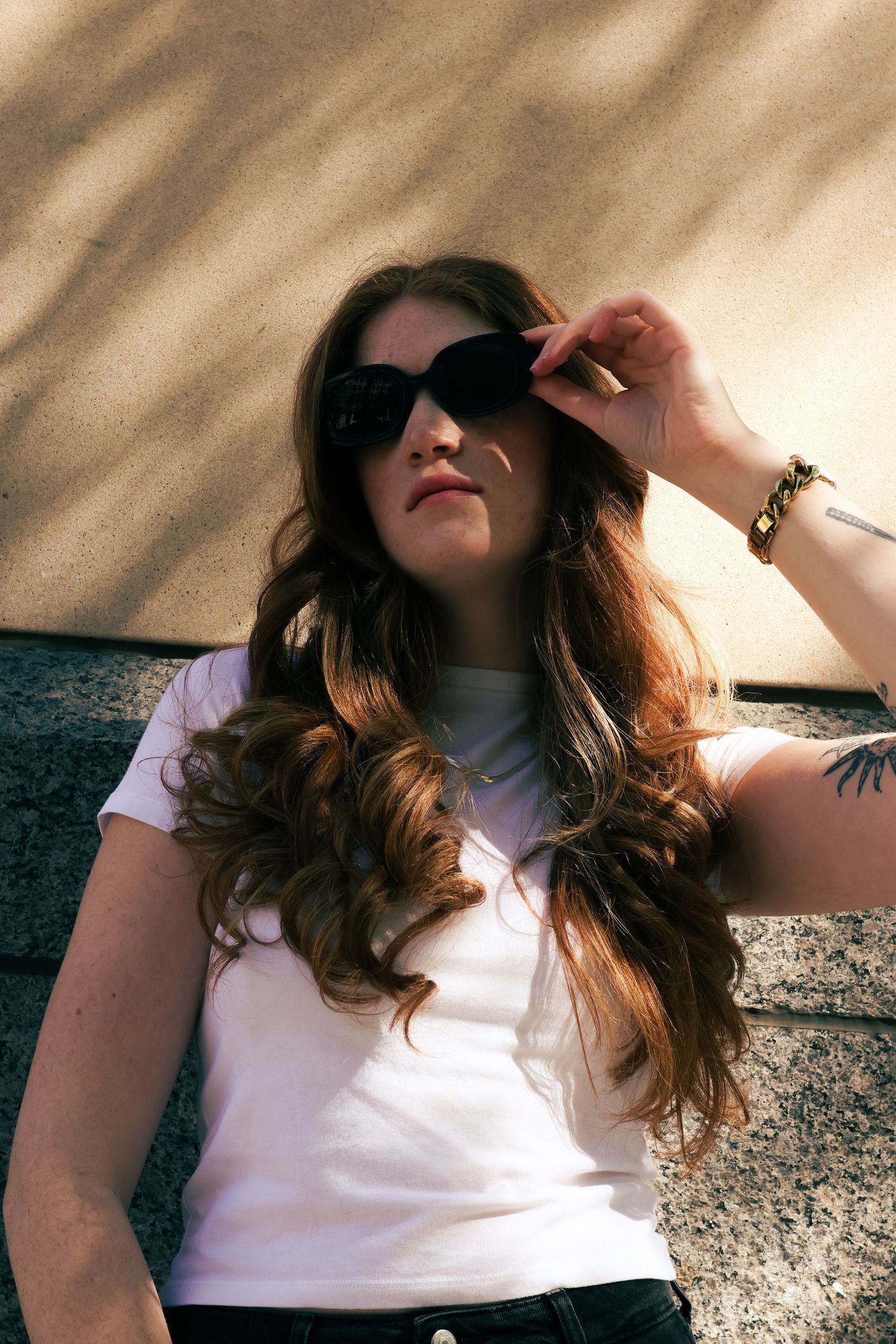 Young woman with long auburn hair wearing sunglasses and white t-shirt with bracelet and arm tattoo poses outdoors.