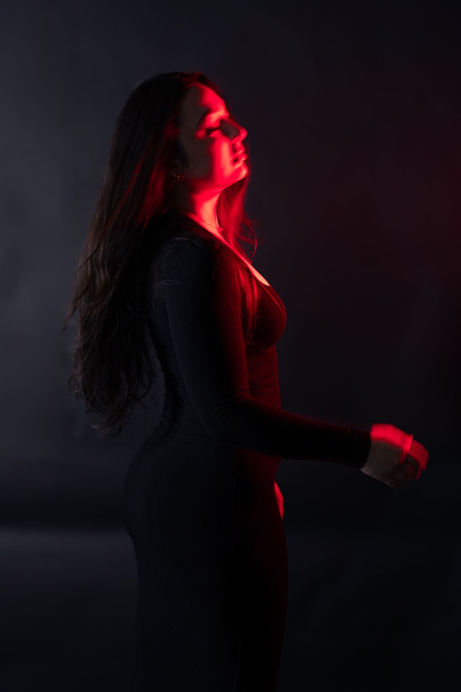 Woman in black outfit posed under red light with smoke in dark studio.