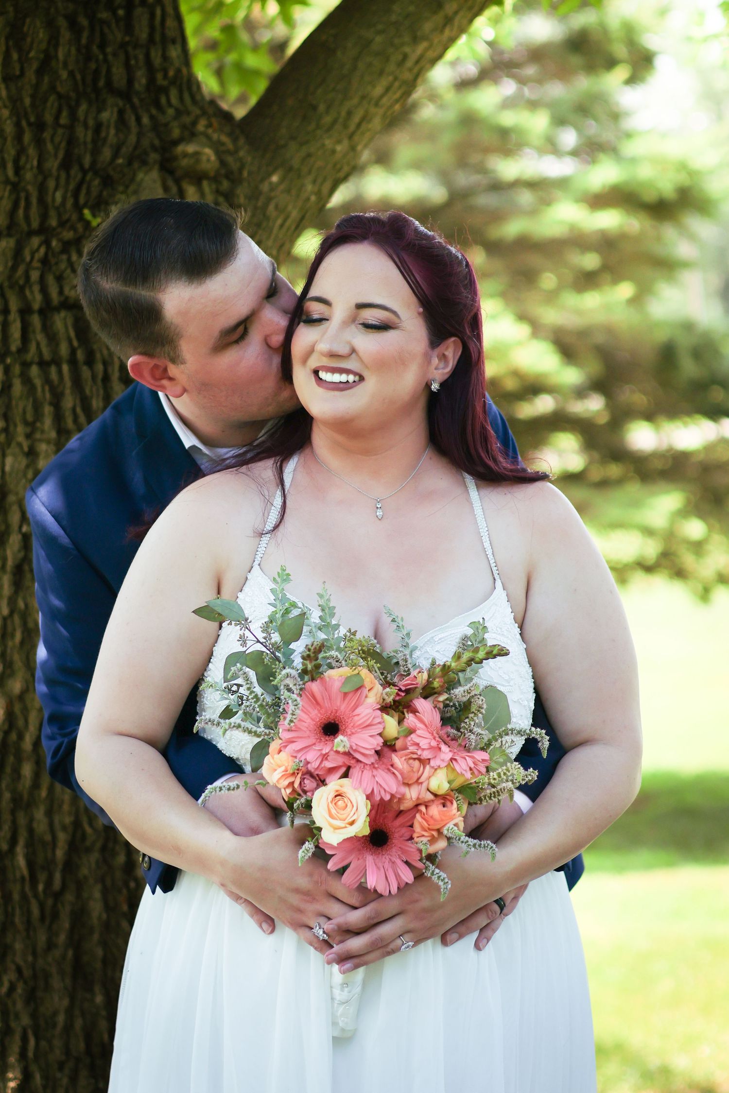 A couple shares an intimate moment by a tree while the bride holds a vibrant pink and coral bouquet.