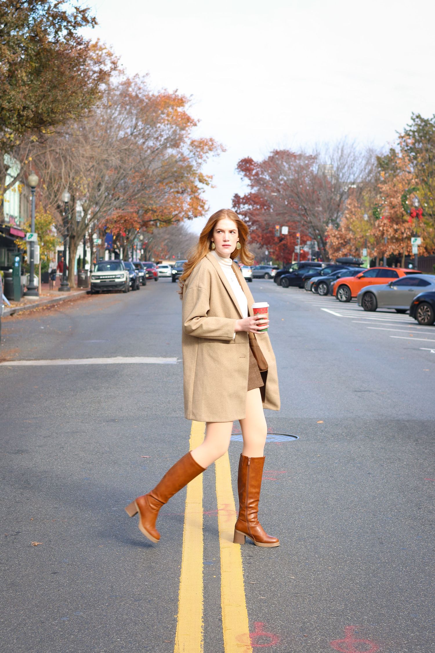 Redhead woman in camel blazer, brown knee boots, and shorts holding red cup while crossing a street in autumn.