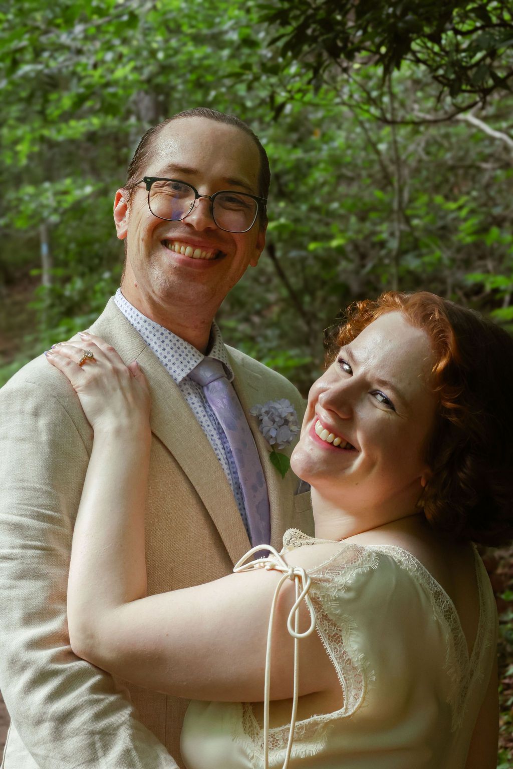 Smiling bride in a cream lace dress embraces a groom in a tan suit with glasses and a boutonniere in a green forest wedding setting.