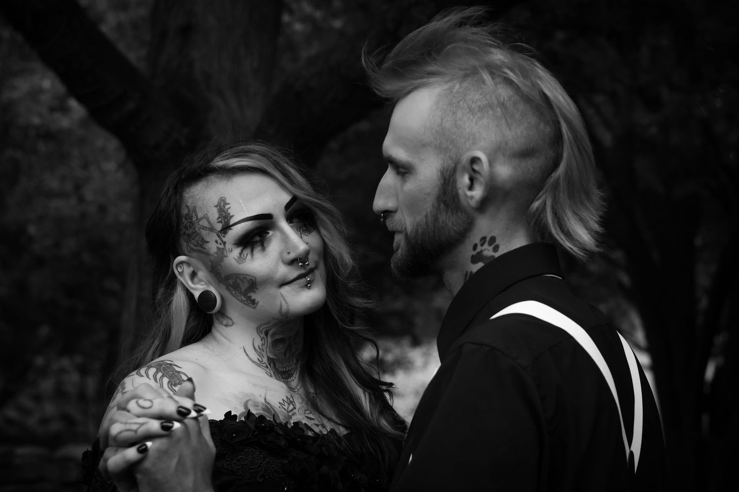 A gothic couple in black and white photography with dramatic makeup and alternative styling pose intimately together.