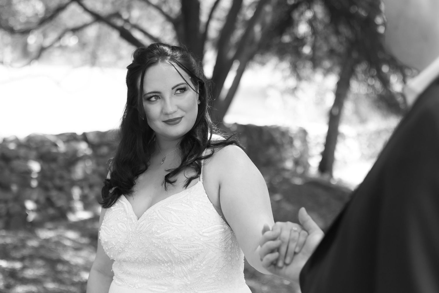 A bride in a white wedding dress poses outdoors among trees in a romantic black and white portrait.