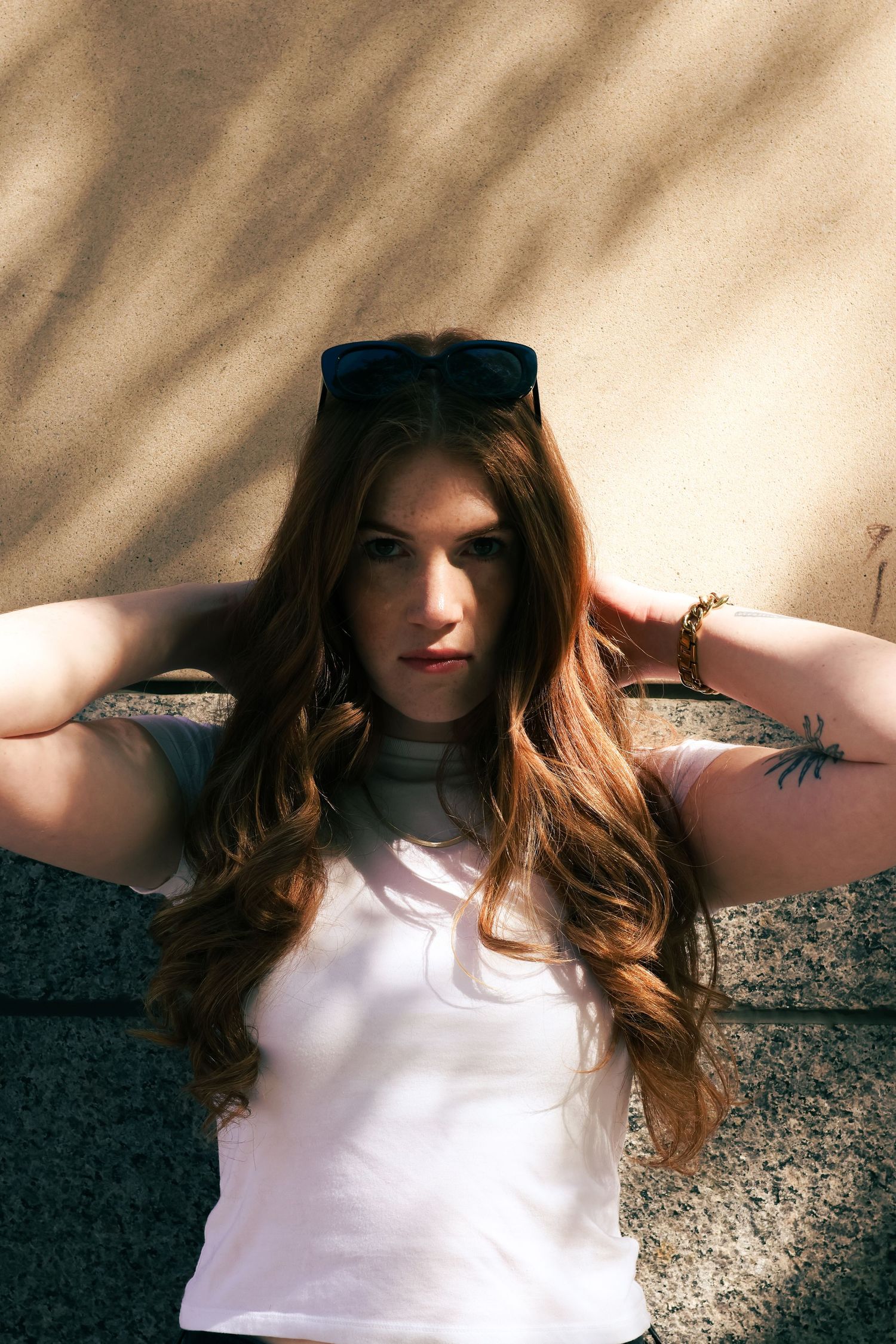 Woman with long wavy auburn hair and tattoo posing against stone wall in white tee.