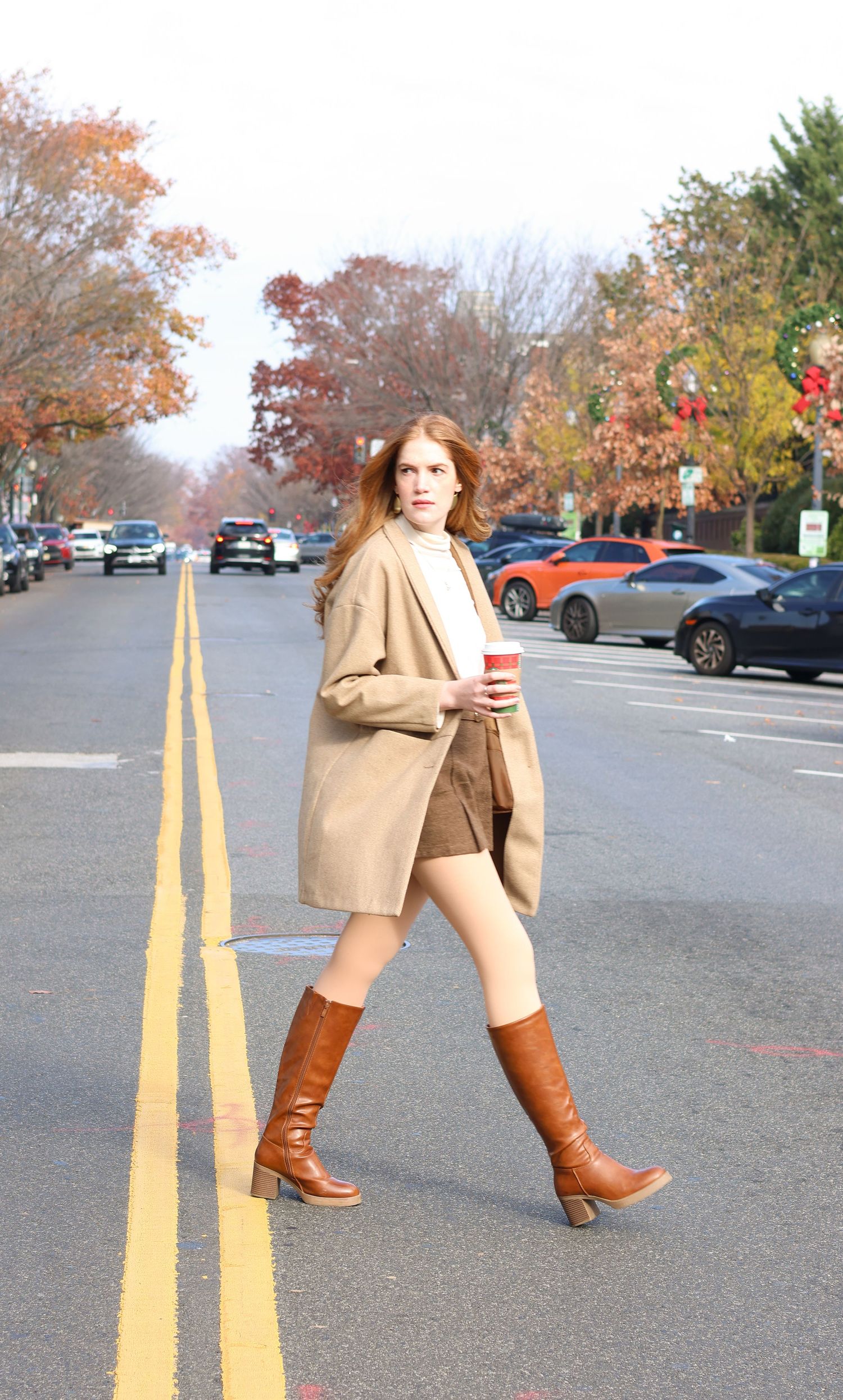 Stylish redhead in camel blazer, brown knee-high boots, and shorts crossing a street holding a red cup in fall.