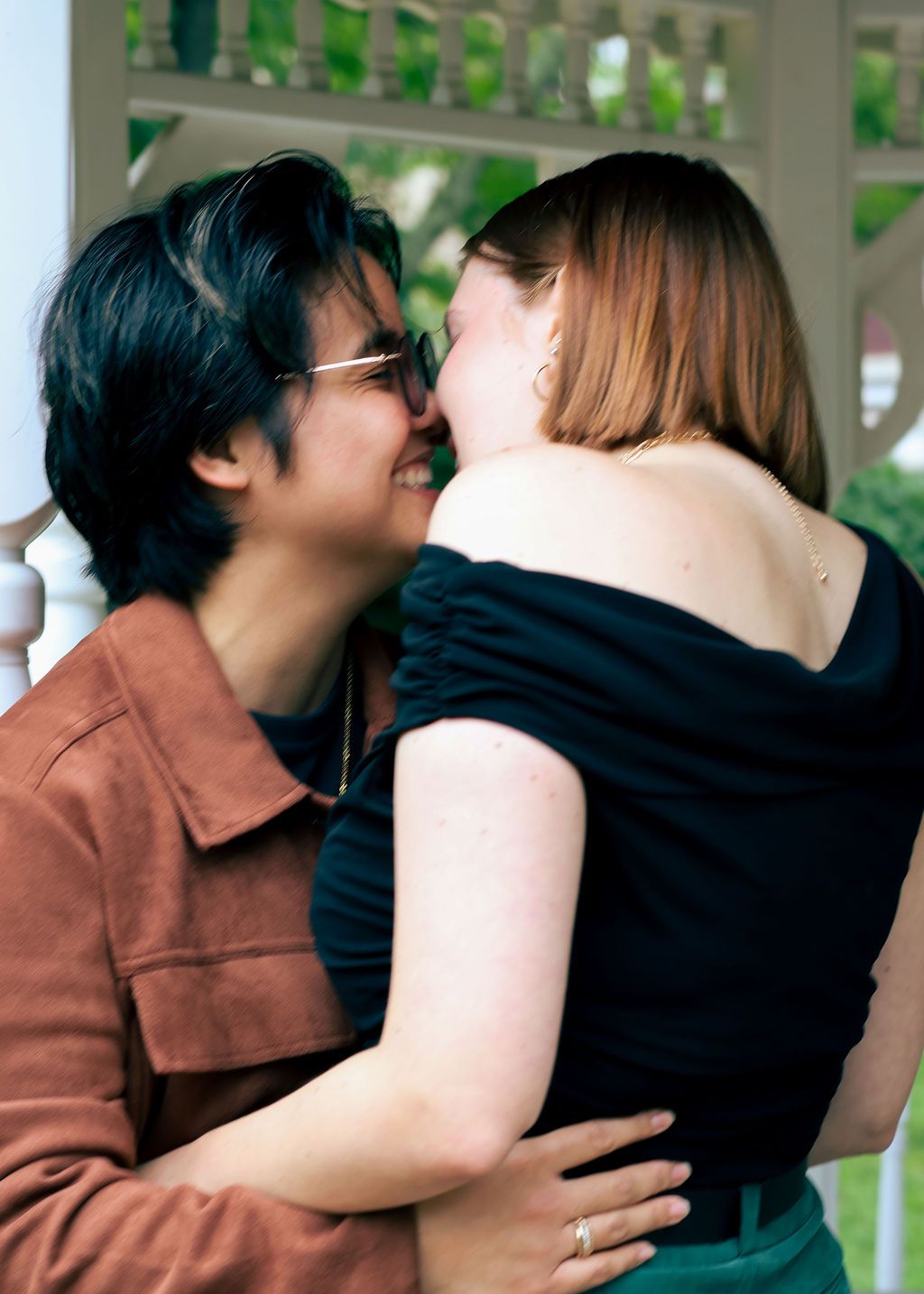 Two people in a warm embrace, one in a rust-colored jacket and glasses, stand close together in a garden gazebo.