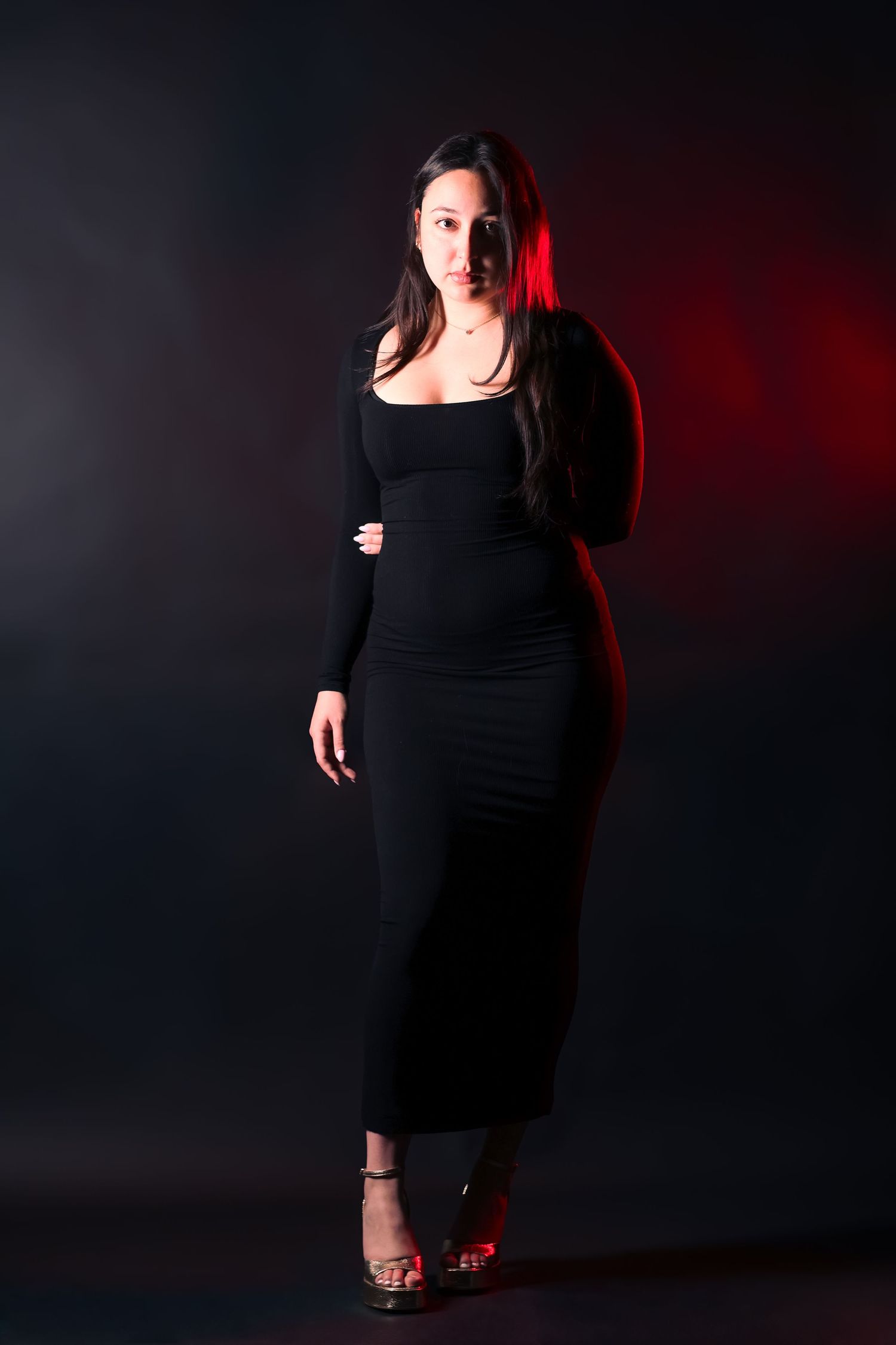 Woman in black long sleeve maxi dress posing confidently with red accent lighting.