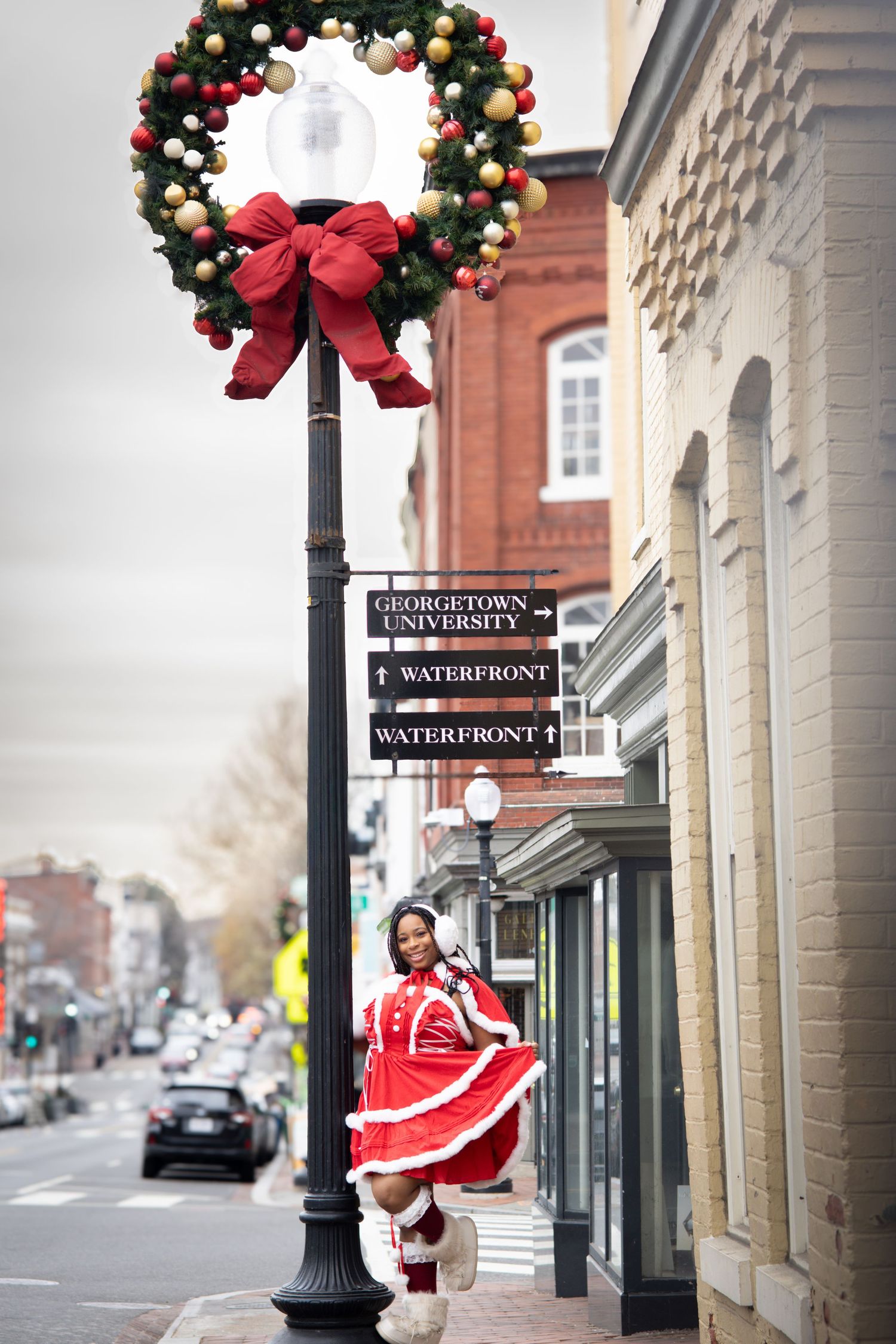 Woman in Santa dress posing by a decorated lamppost with Christmas wreath near Georgetown University waterfront sign.