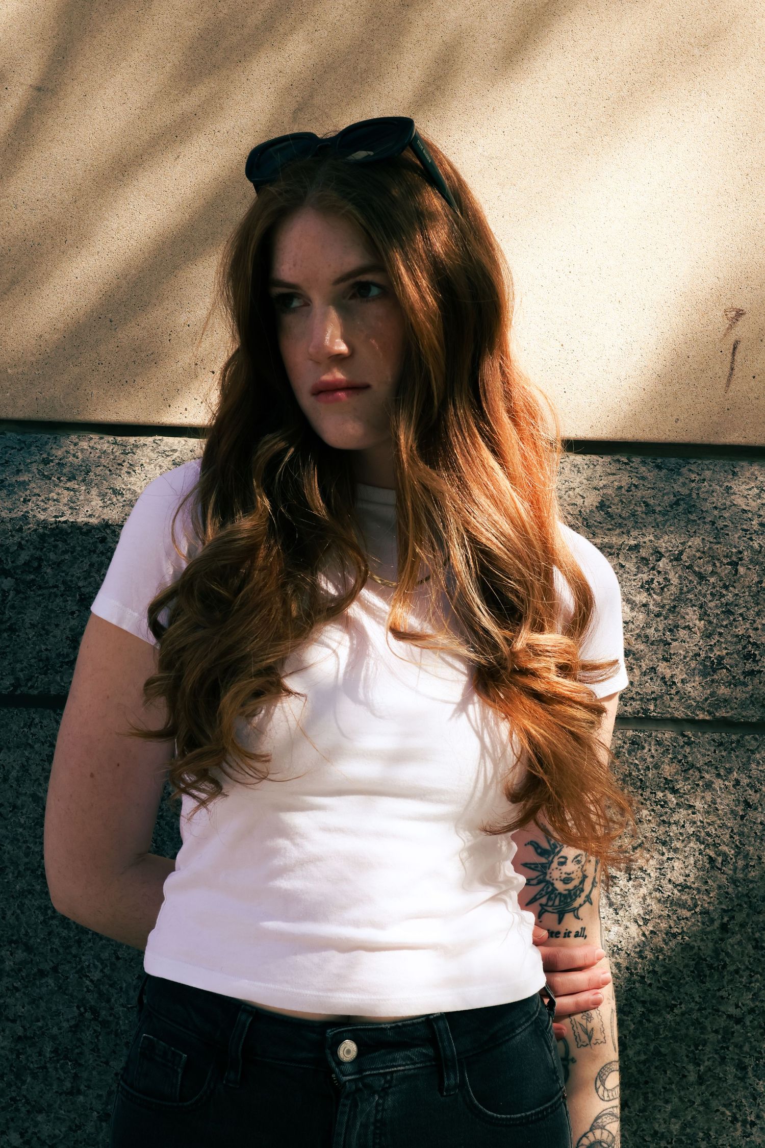 Young woman with long auburn hair, freckles, white t-shirt, sunglasses on head, and arm tattoo poses outdoors.